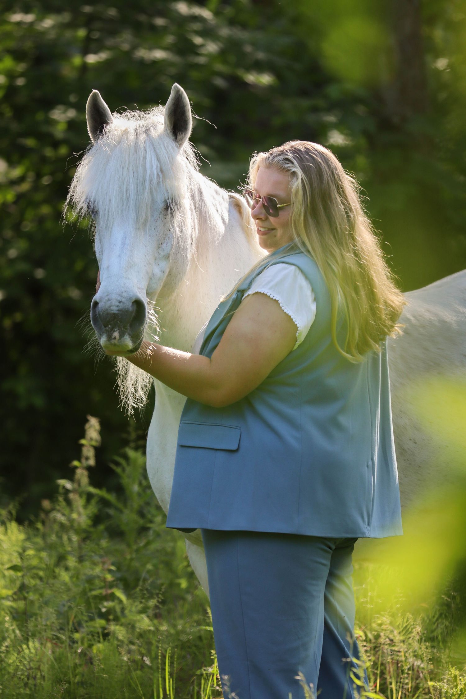 Blonde woman in blue vest tenderly bonding with a white horse in a sunlit green forest.