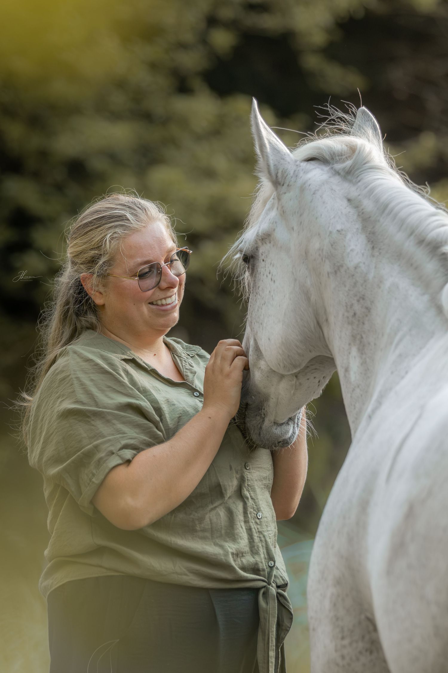 A smiling woman with glasses pets a white horse outdoors, sharing a joyful moment against a green background.