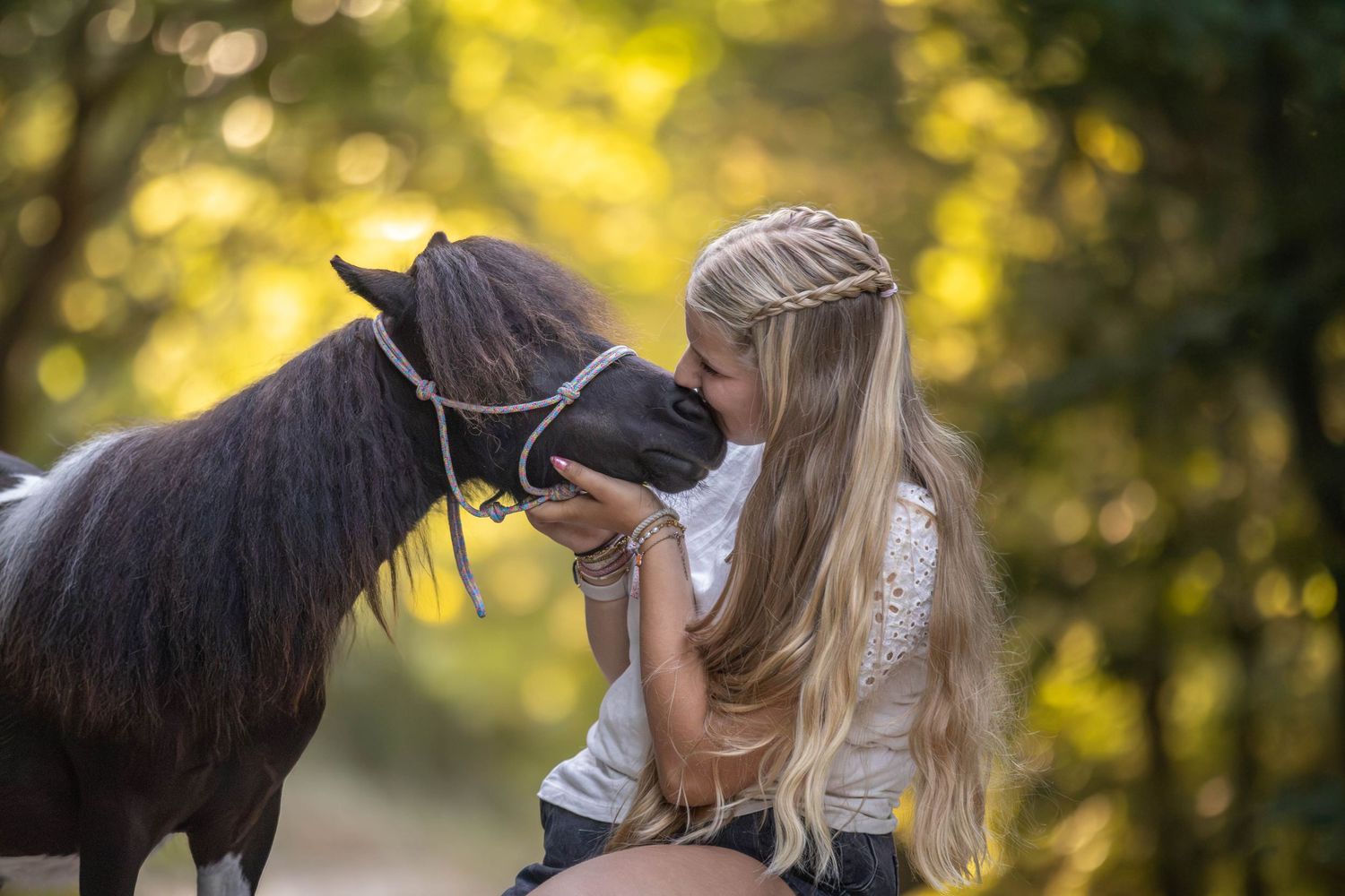 Young blonde girl kissing a small dark pony on a sunlit forest path.