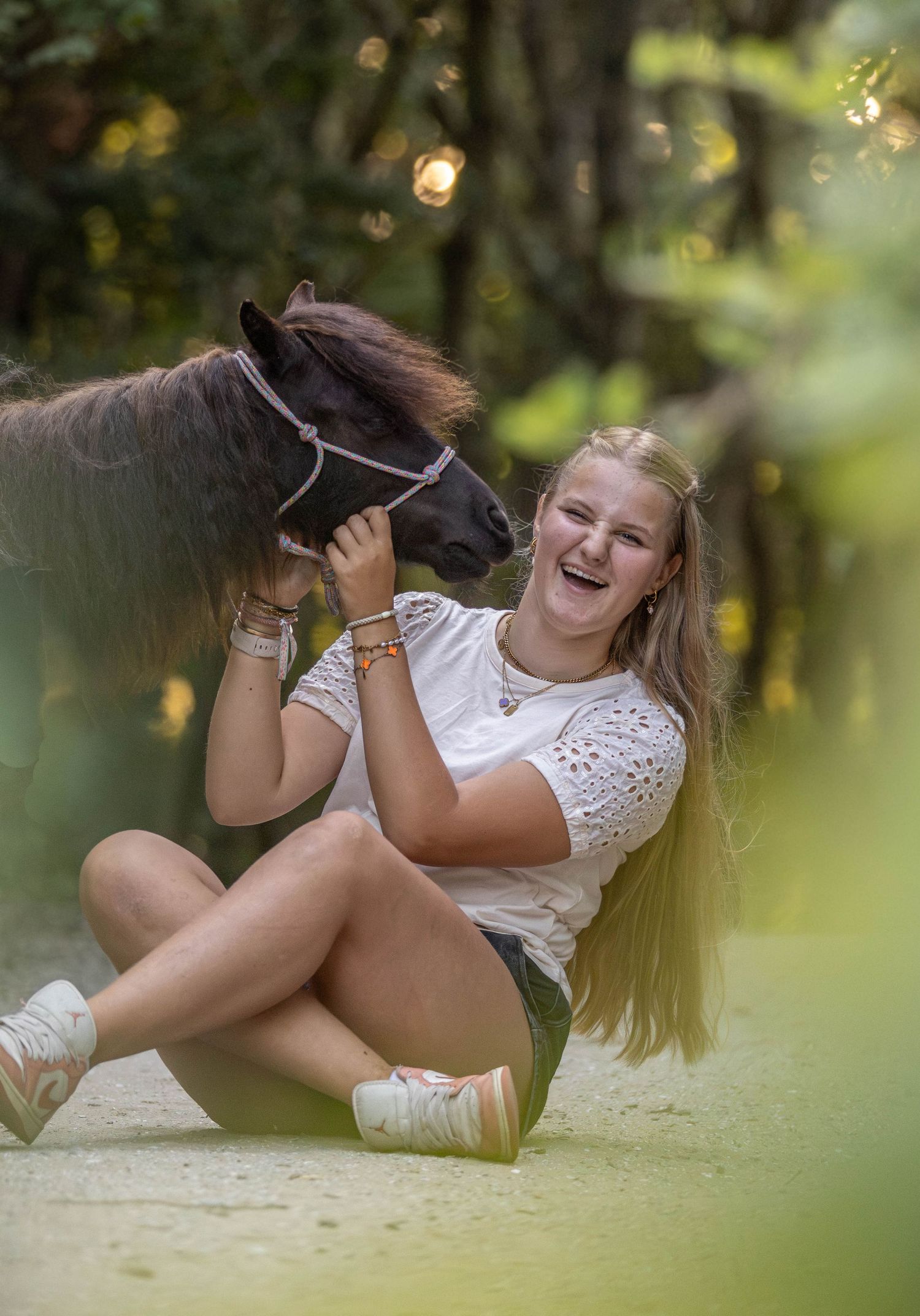 Laughing blonde girl sitting on a sandy forest path cuddling a small dark pony wearing a halter.