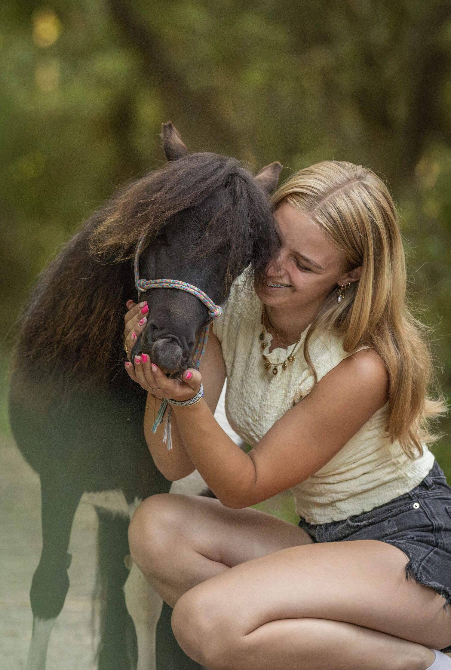 Smiling blonde woman crouching outdoors, affectionately hugging a small dark pony in a forest setting.