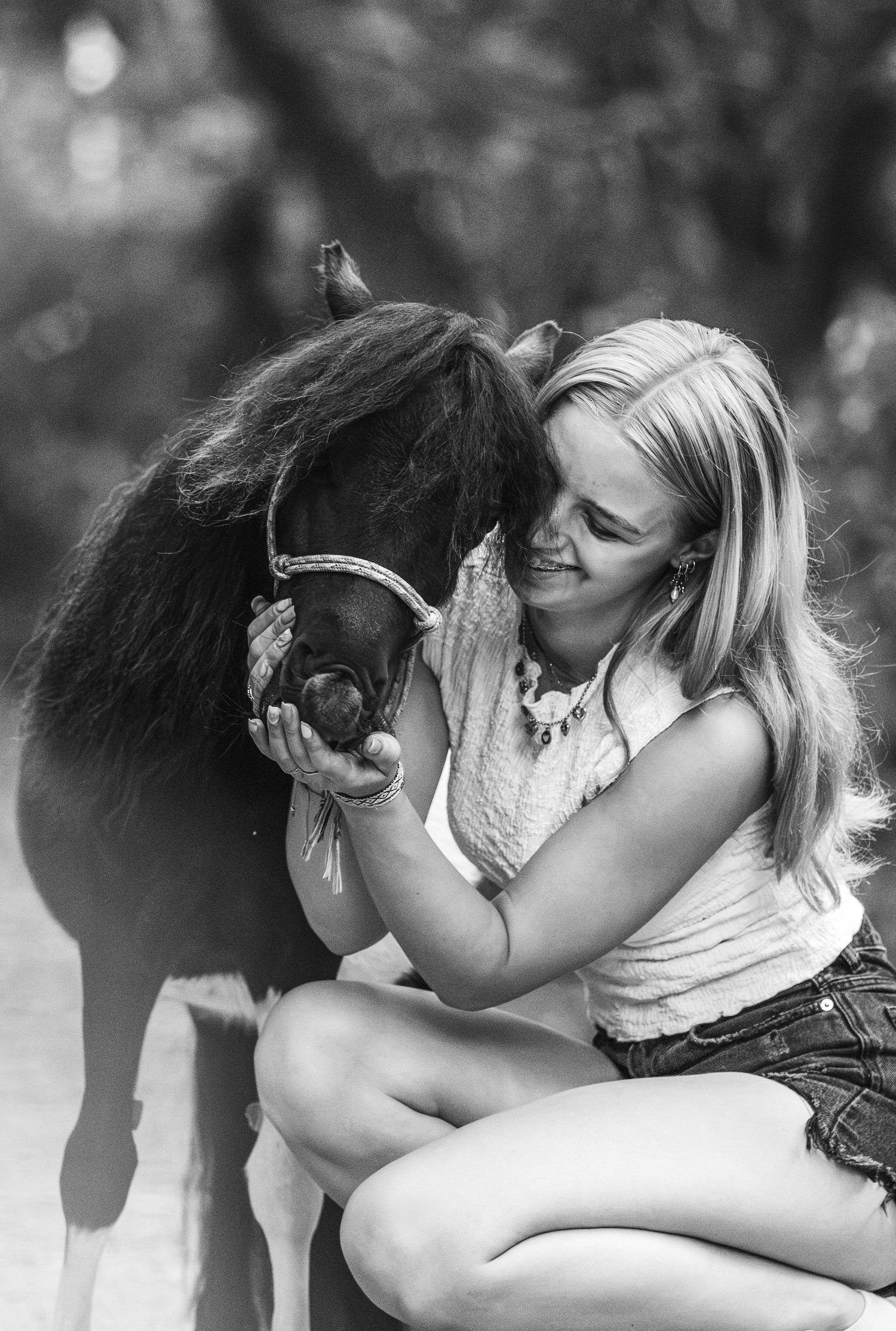 A blonde girl embraces a small dark pony affectionately in this tender black and white outdoor portrait.