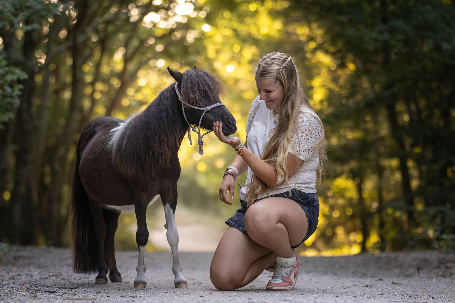 Young blonde woman kneeling on a dirt path feeding or petting a small dark pony with a halter in a forest.