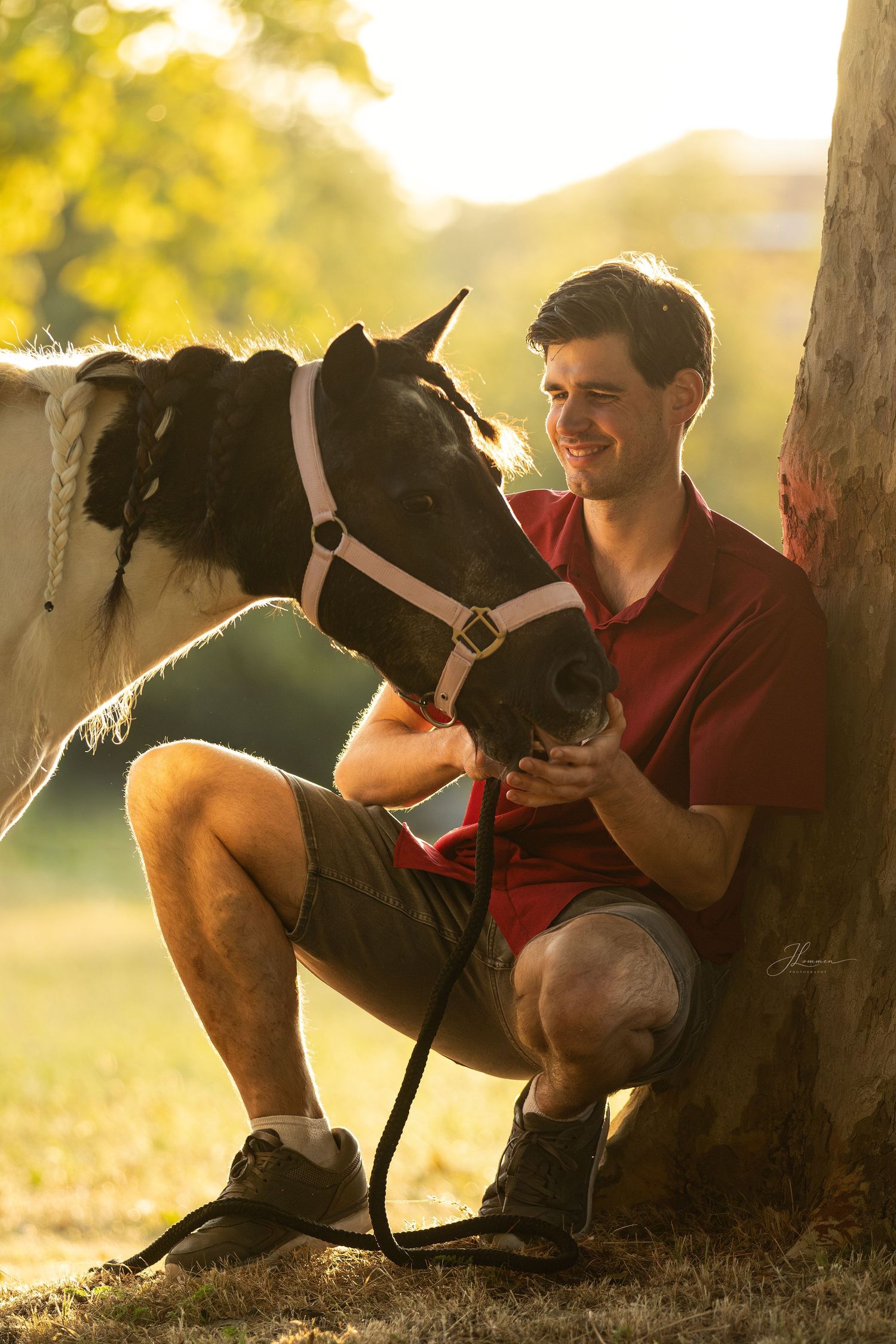 Smiling man in red polo shirt crouching beside a black and white horse near a tree in golden sunlight.