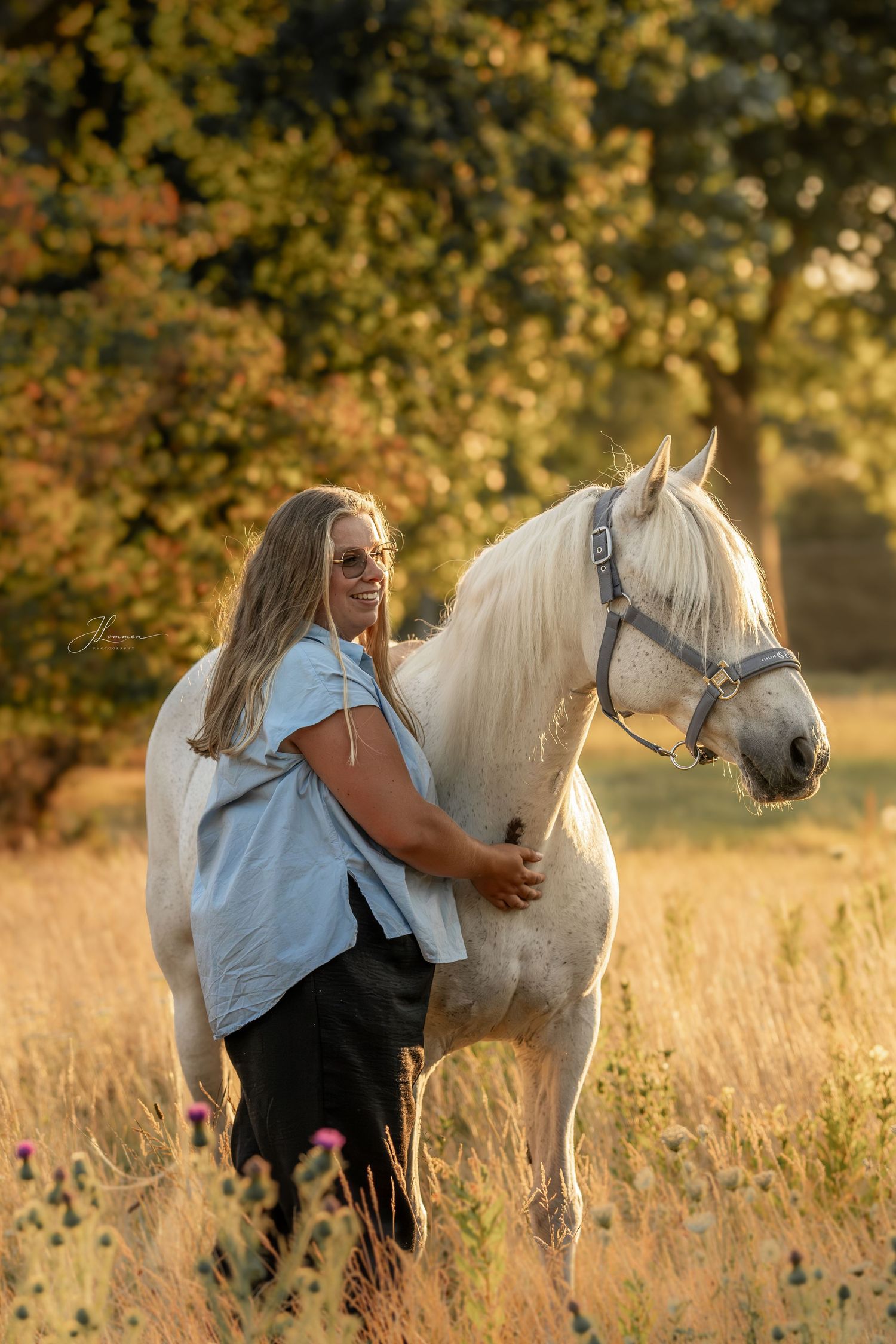 A smiling woman in a blue shirt stands beside a white horse in a sunlit golden field with autumn trees.