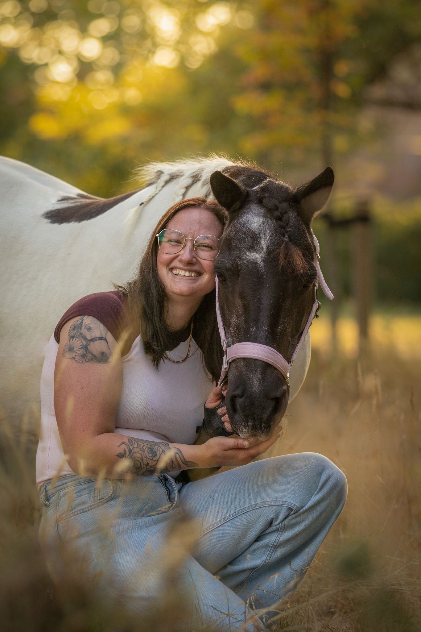 A smiling woman with glasses and tattoos crouches beside a black and white horse in a sunlit autumn field.
