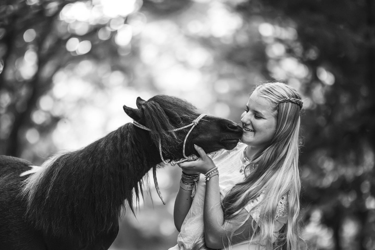 Blonde girl smiling while a small pony nuzzles her face, black and white photo.