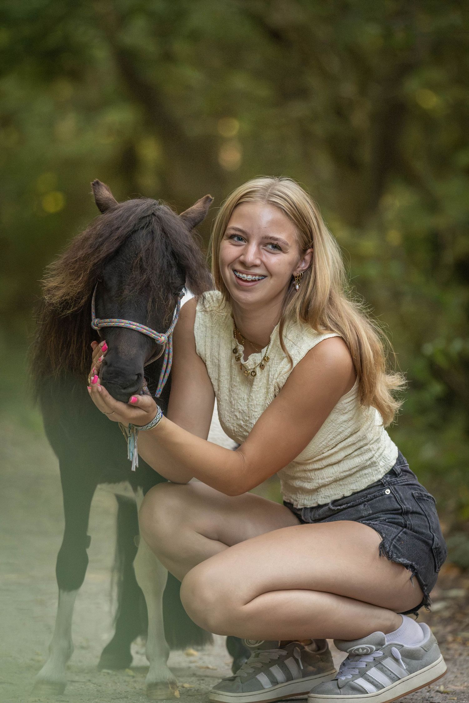 A smiling blonde teenage girl crouches beside a small dark pony on a forest path, hugging its head warmly.