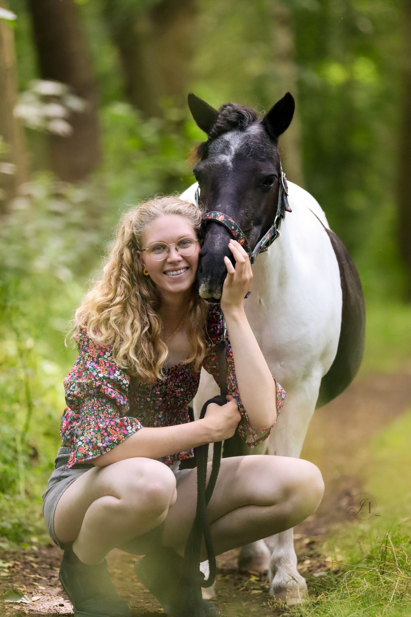 Smiling blonde woman with glasses kneeling beside a black and white horse in a green forest.