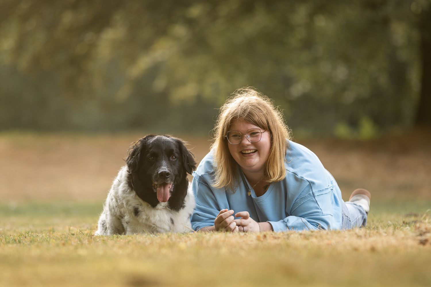 Woman with glasses lying on grass smiling beside a large black and white dog in a sunny park.