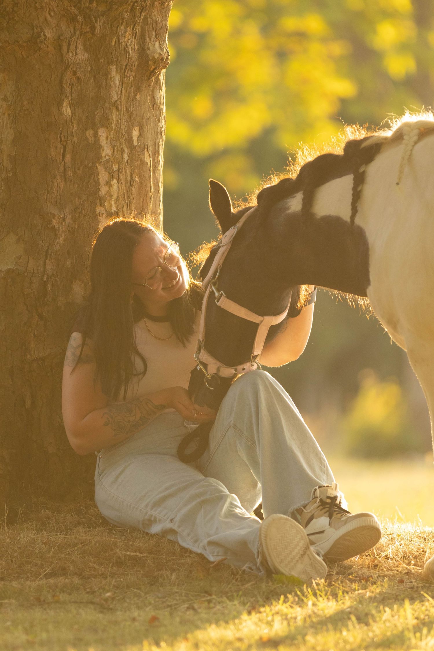 Young woman sitting under a tree in golden light sharing a tender moment with a pinto pony.