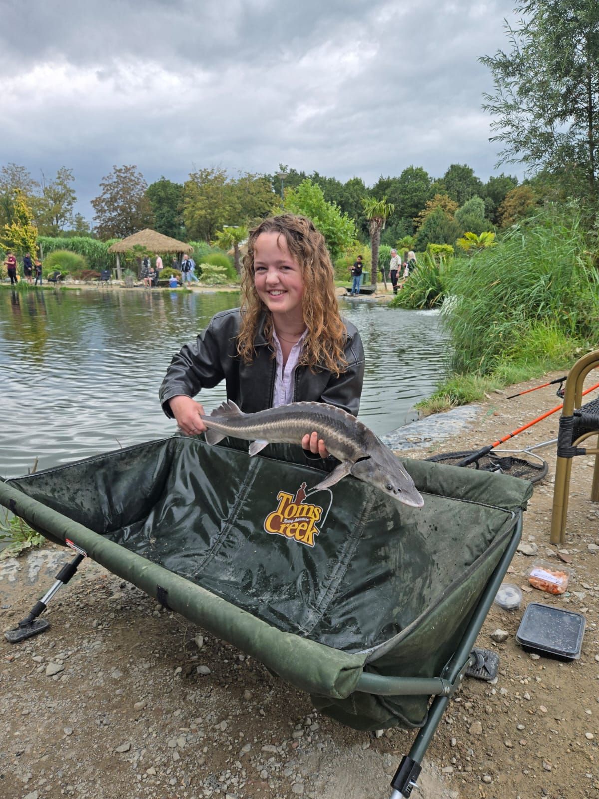 Young girl smiling while holding a large fish over a green Tom's Creek fishing mat beside a scenic lake.