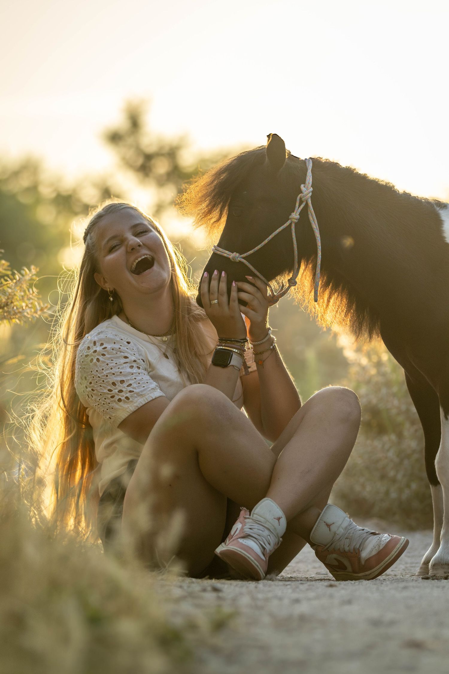 Laughing blonde girl sitting on ground with small dark pony in golden sunset light.