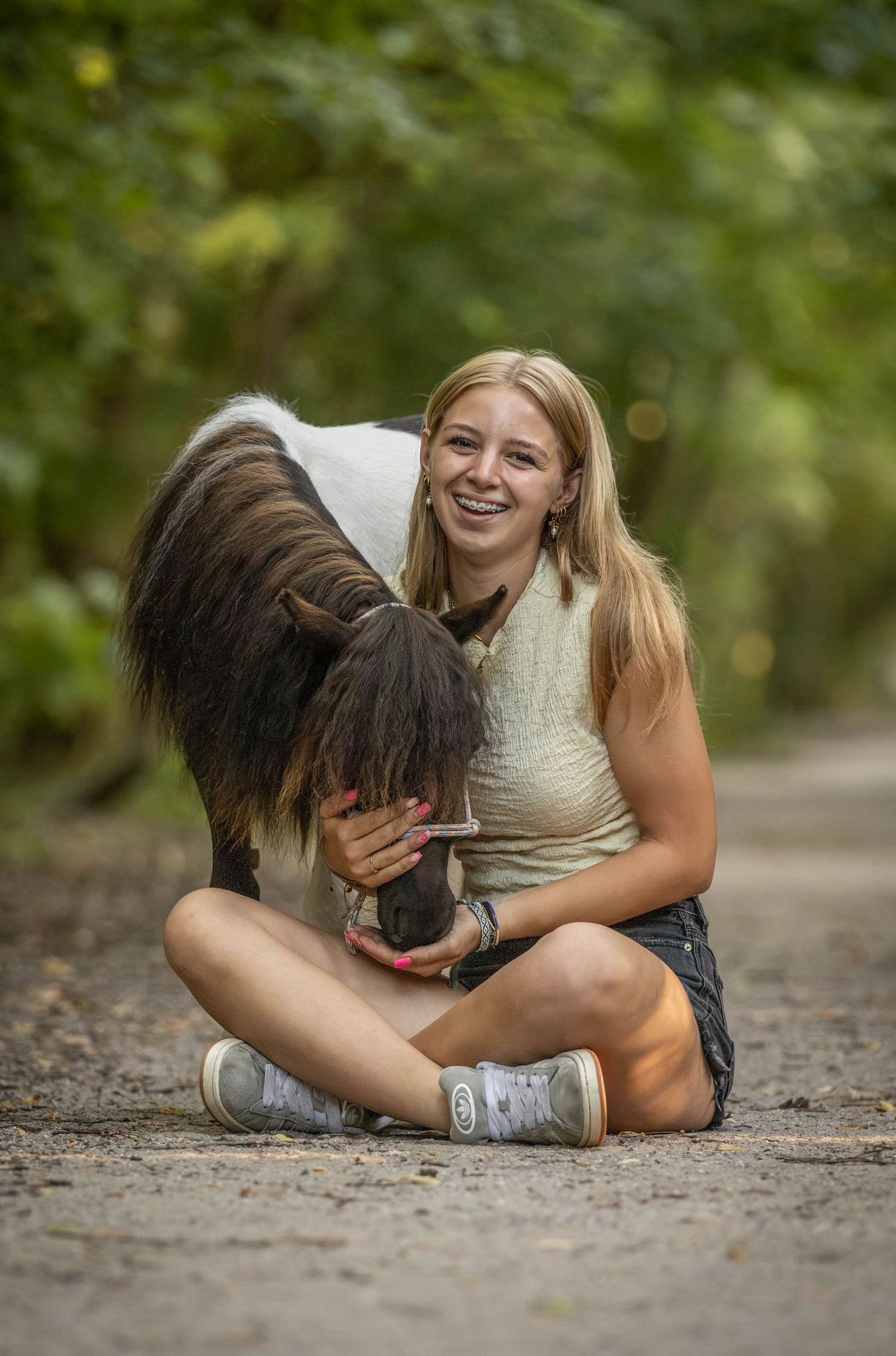 Smiling blonde girl sitting cross-legged on a path hugging a small black and white pony in a green forest.