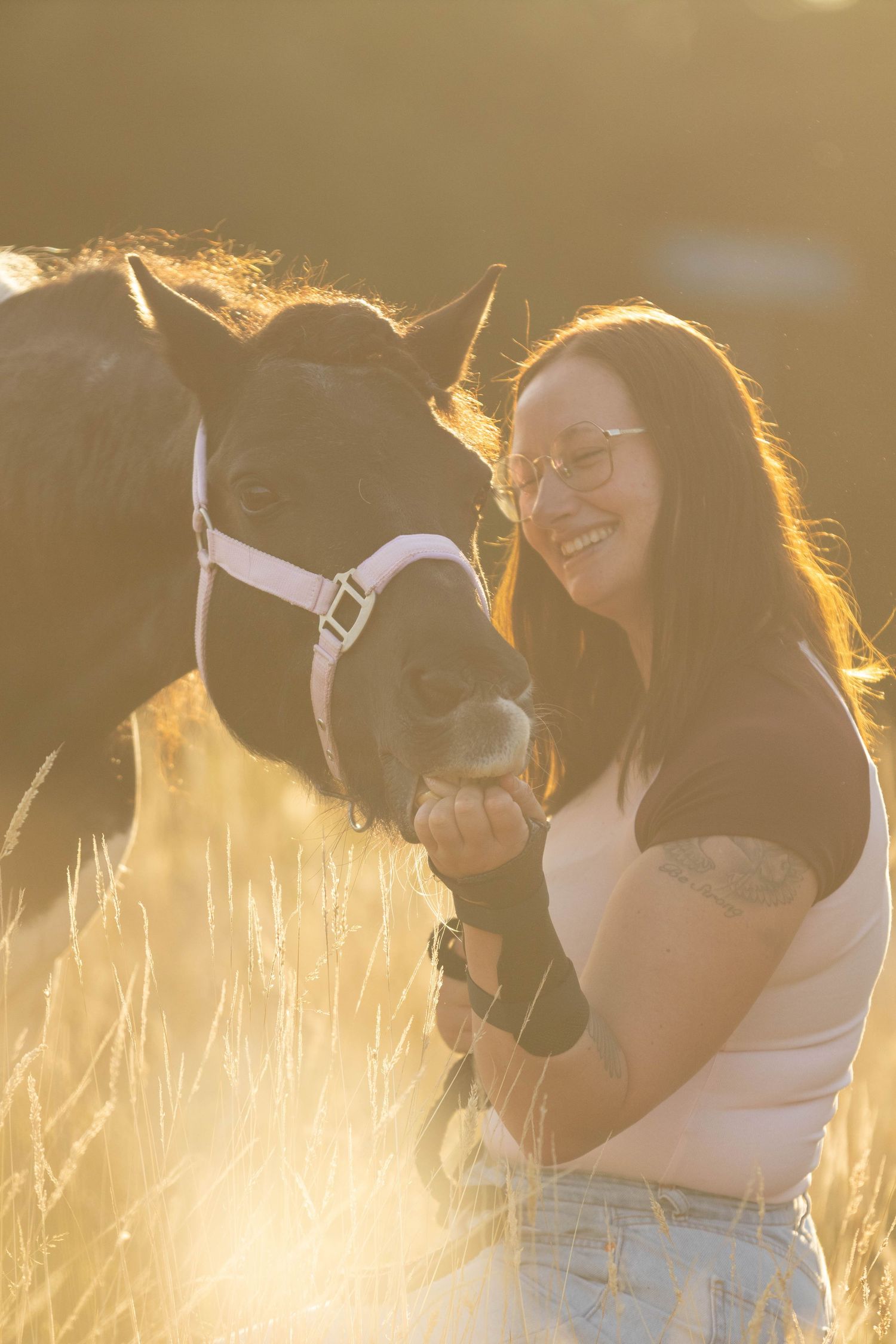 Smiling brunette woman with glasses feeding a dark pony by hand in a golden sunlit field.