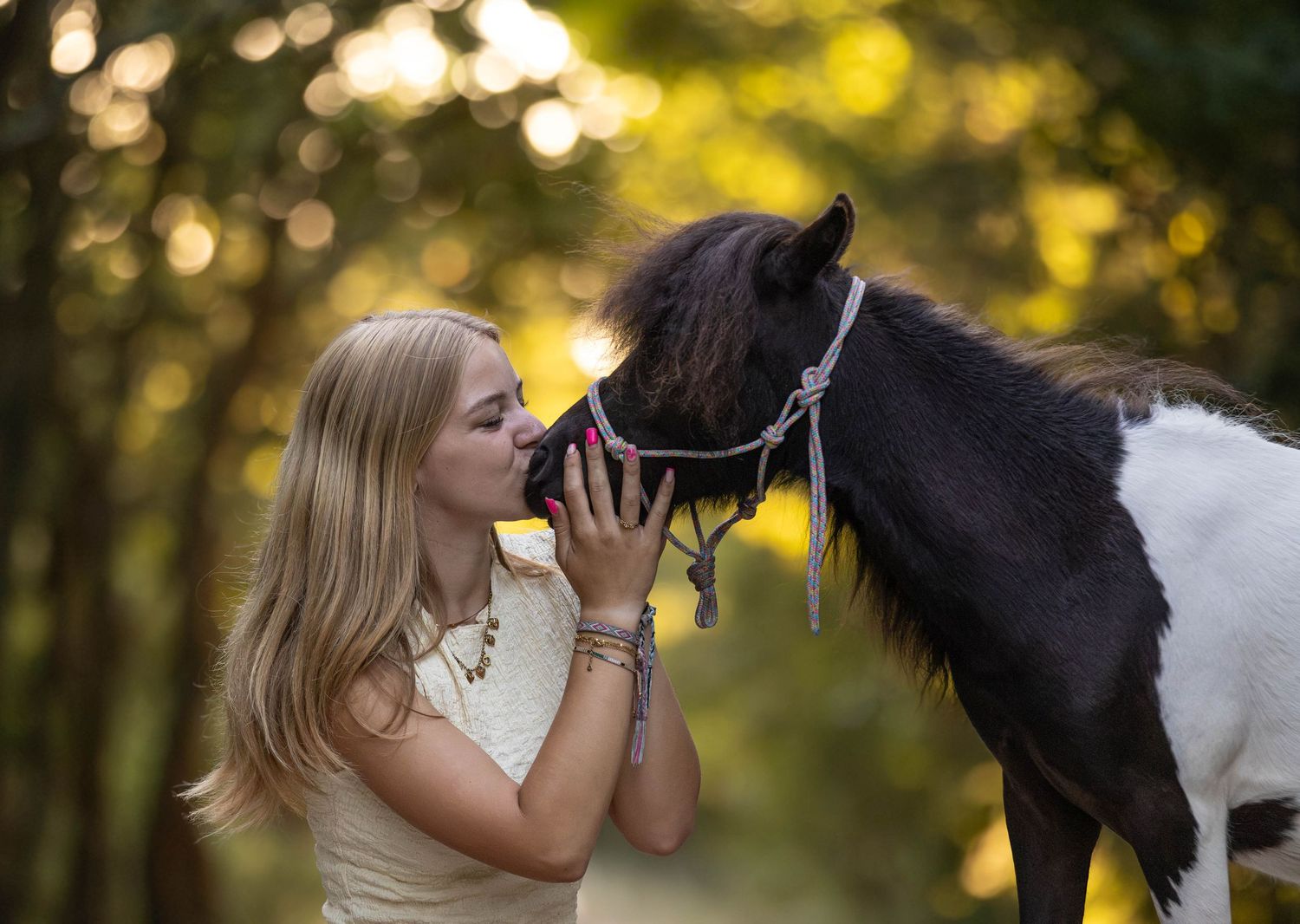 Young blonde woman kissing the nose of a small black and white pony wearing a pink halter in a forest.