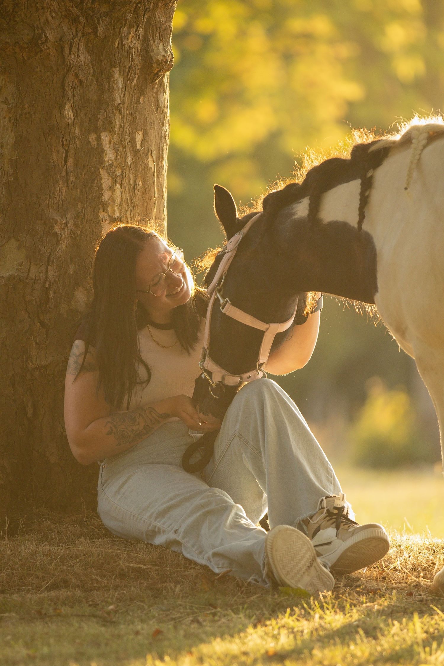 Young woman sitting against a tree in golden sunlight, smiling while gently petting a black and white horse.