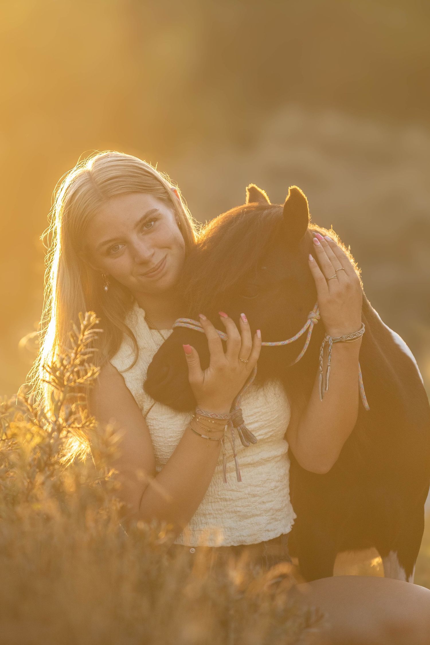 Smiling blonde girl hugging a small brown pony in a golden sunset field.