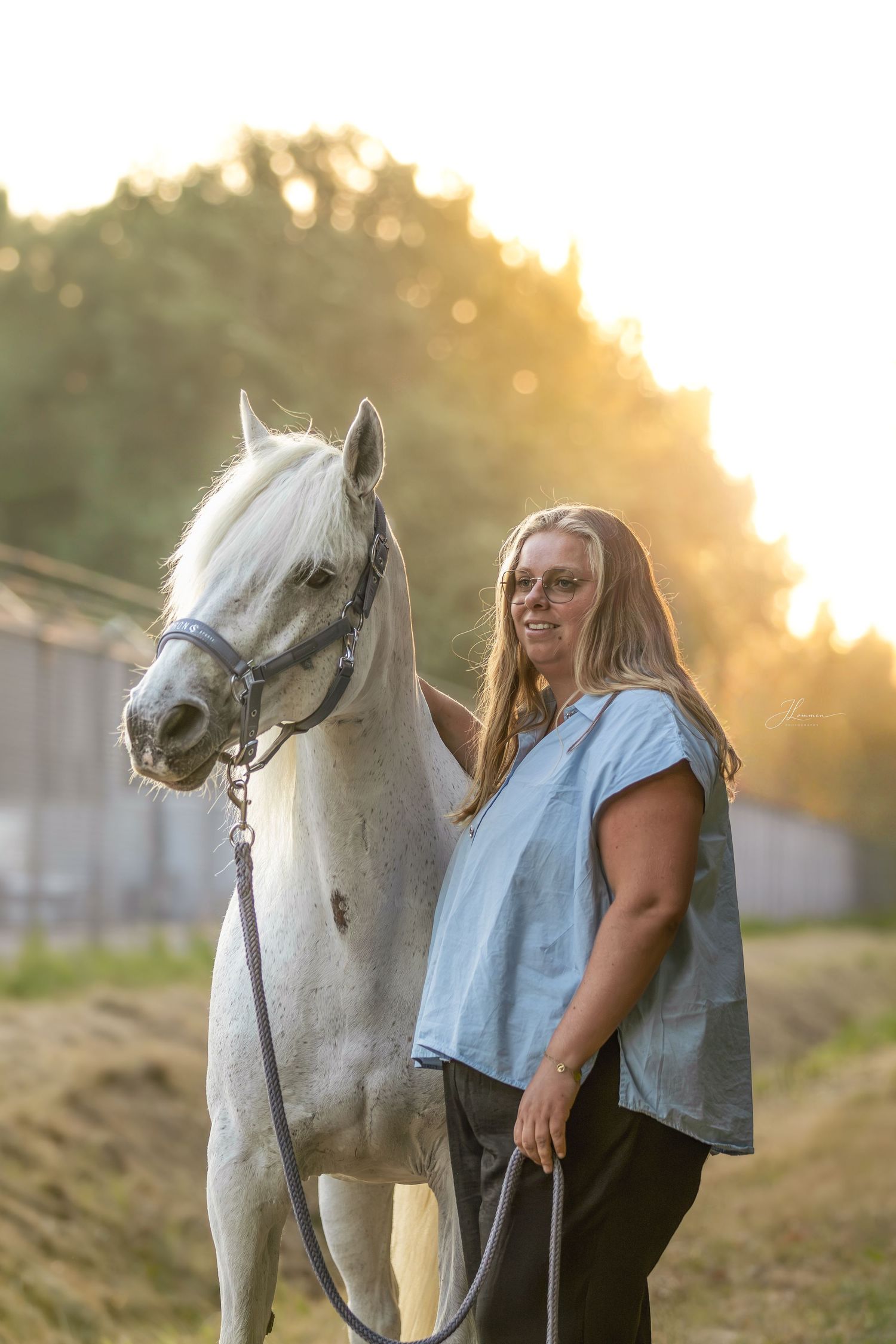 Woman in blue top smiling beside a white horse at golden hour outdoors.