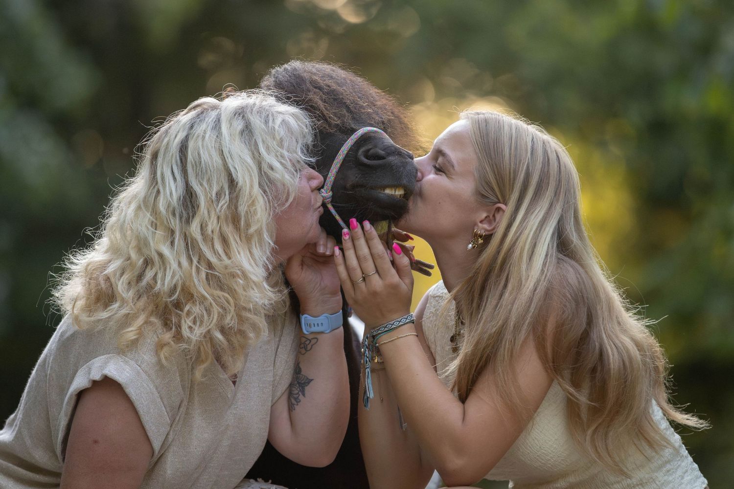 Two blonde women both kissing a small dark pony on the nose outdoors in a sun-dappled garden setting.