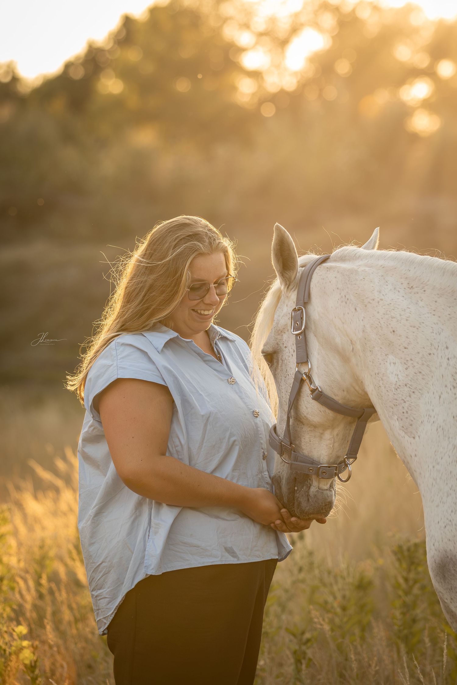 Woman with blonde hair smiling while holding a white horse in a golden sunset field.