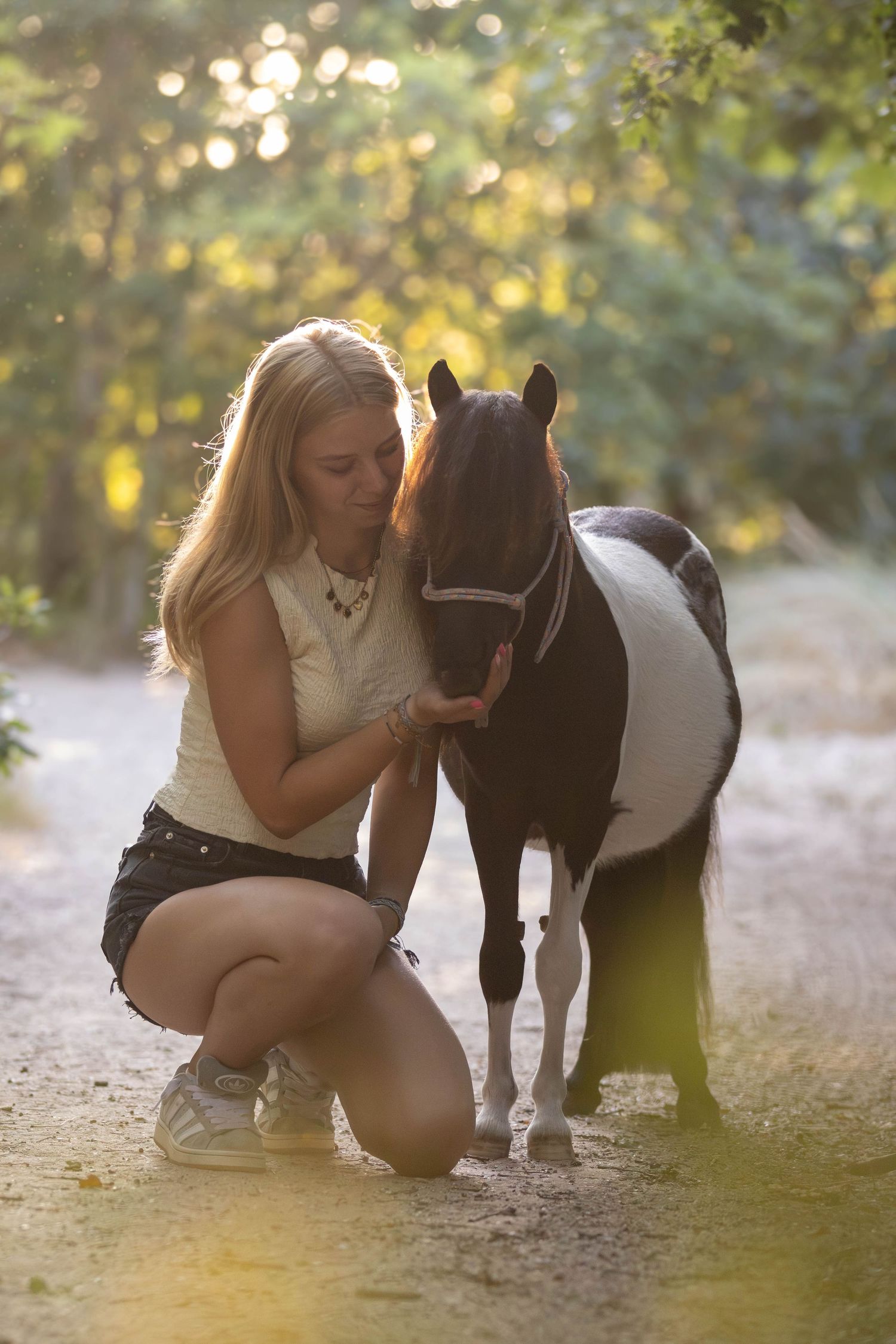Blonde girl kneeling beside a small black and white pony on a sunlit forest trail.