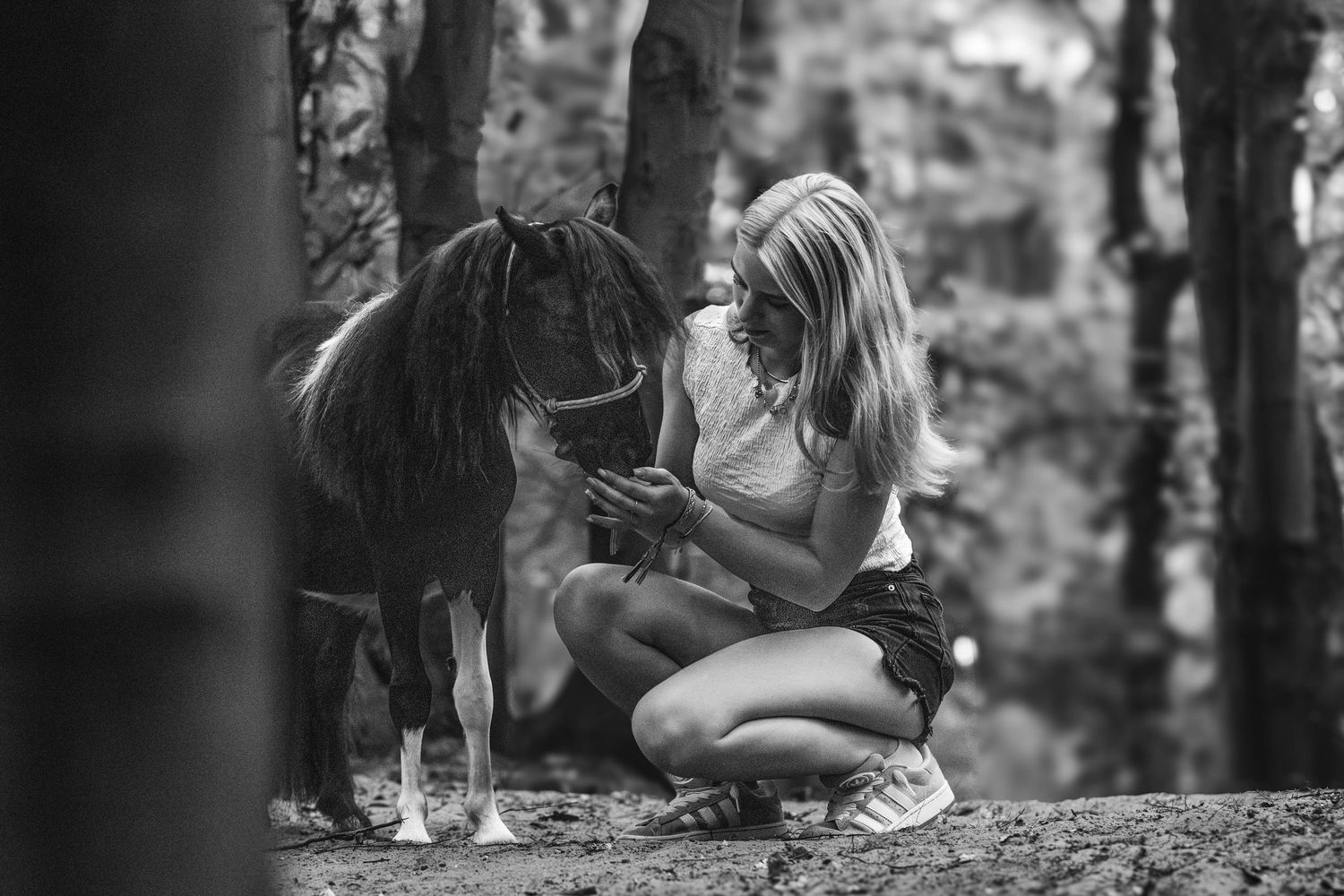 Black and white photo of a blonde girl crouching beside a small pony in a forest, looking at her phone.