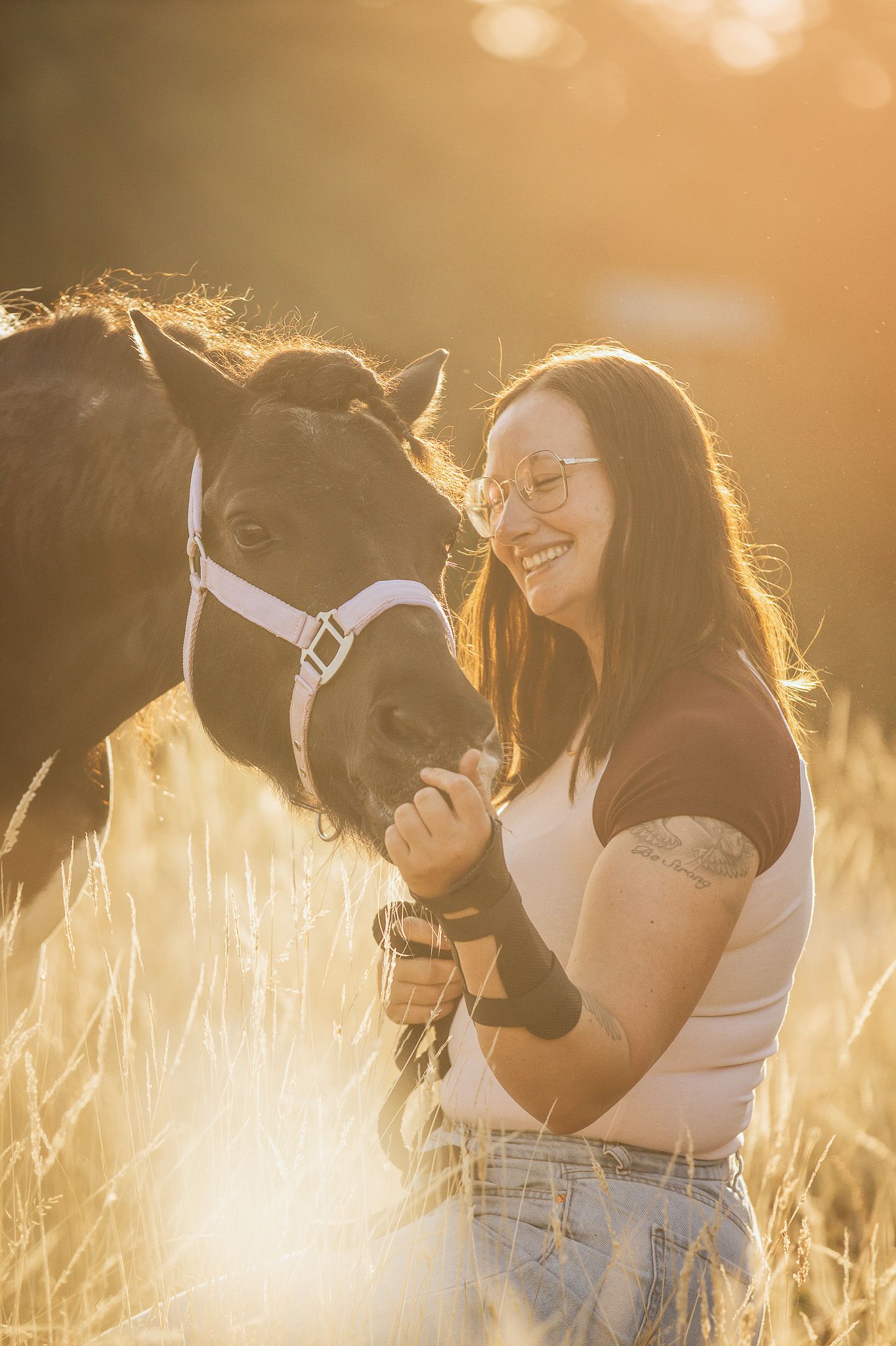 Woman smiling with a dark horse in golden sunlight field, wearing glasses and a black top with visible tattoo.