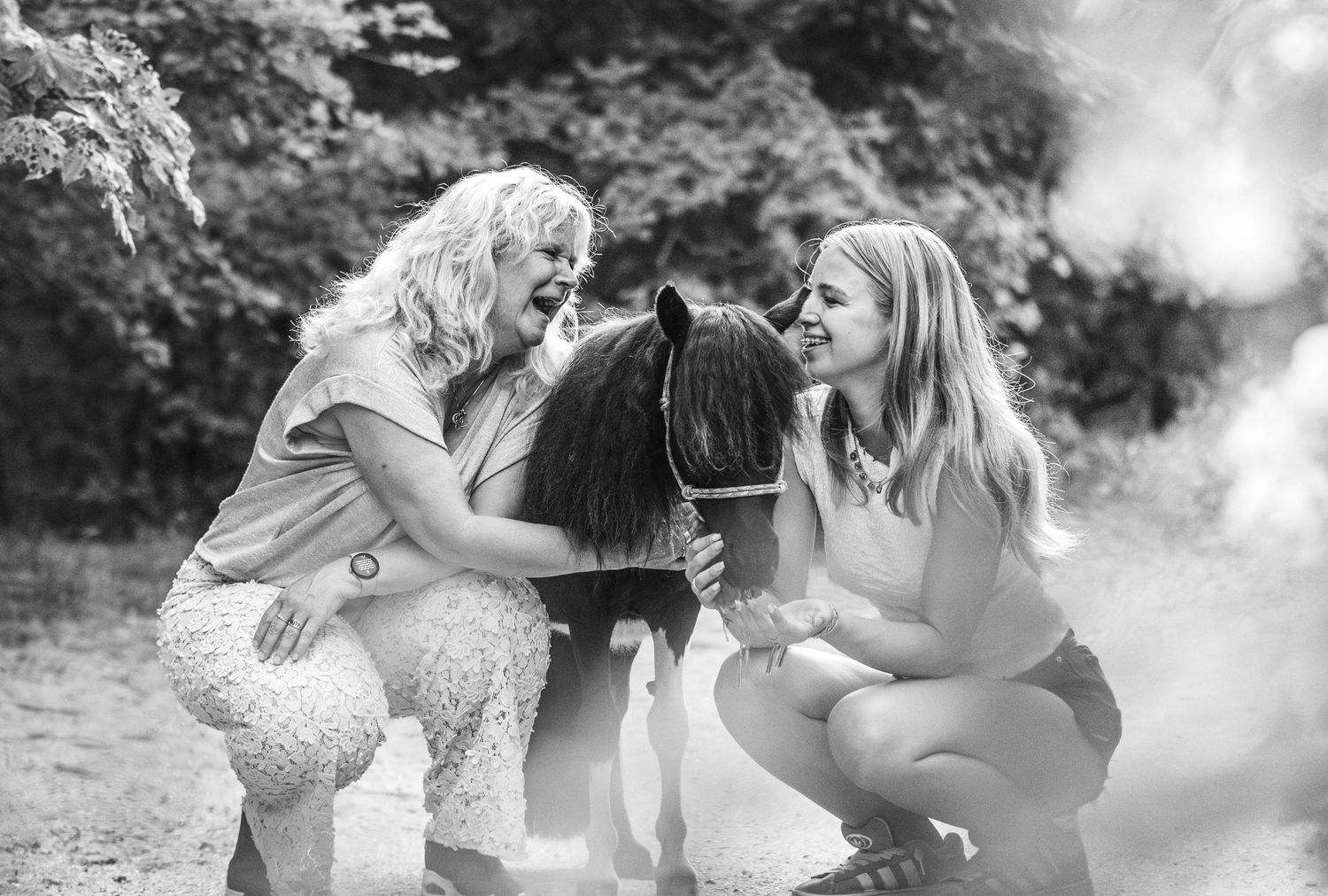 Black and white photo of two women laughing and petting a small dark fluffy pony outdoors.