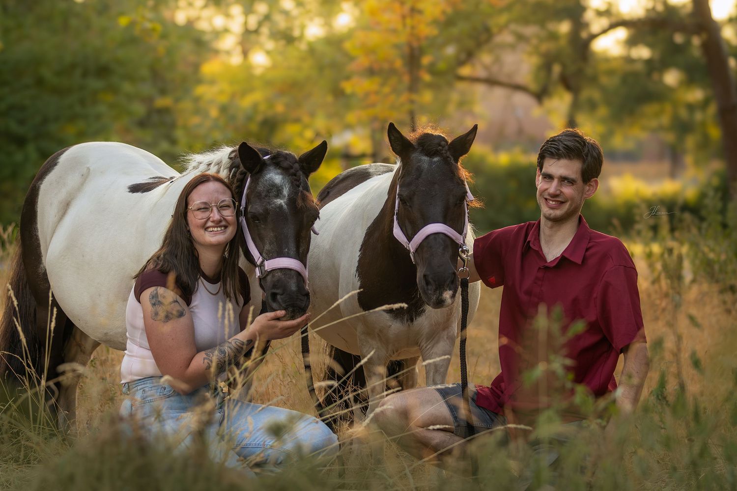 A smiling couple sits in a golden meadow with two paint horses behind them during a warm sunset.