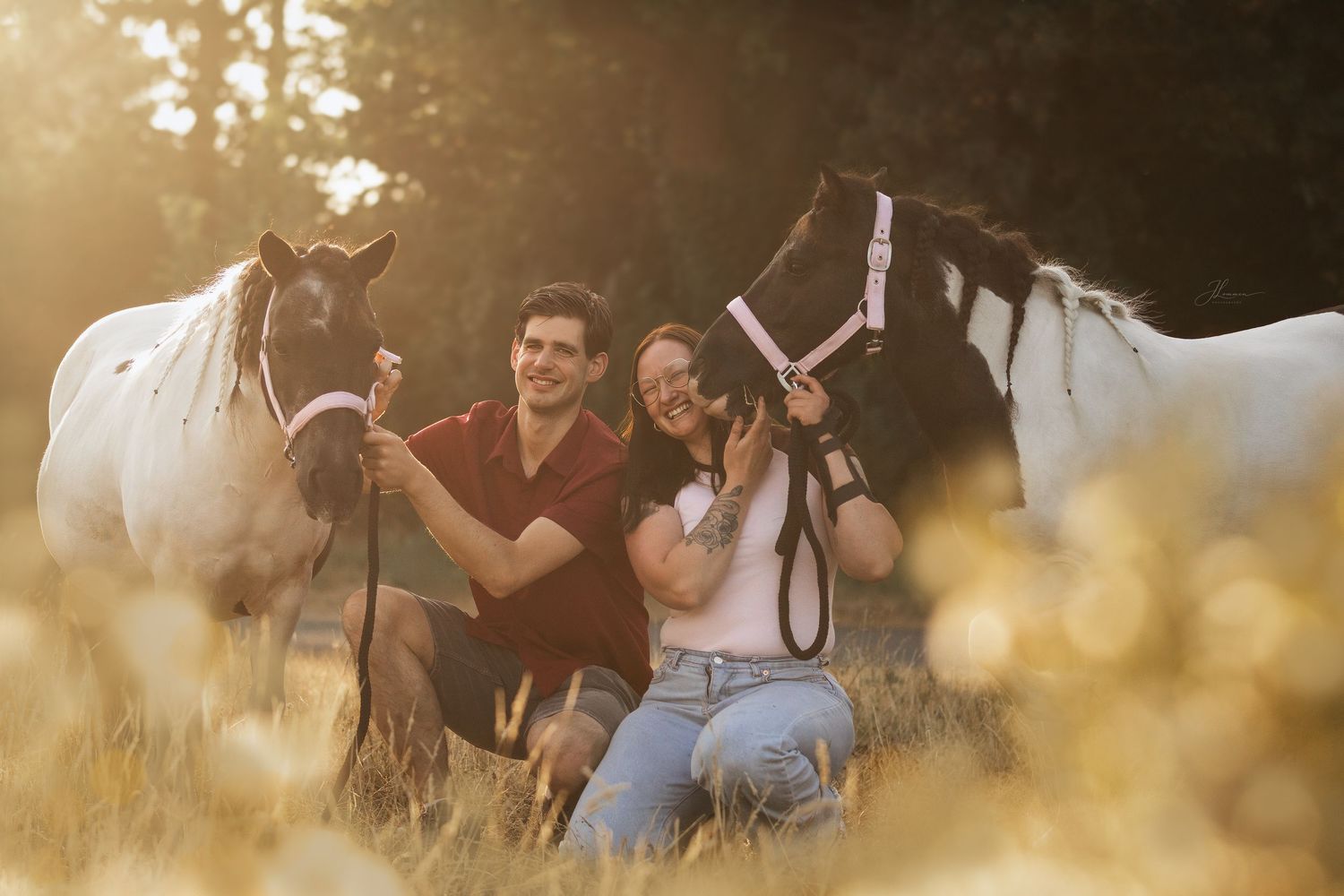 Couple sitting in golden field with two horses in warm sunset light.