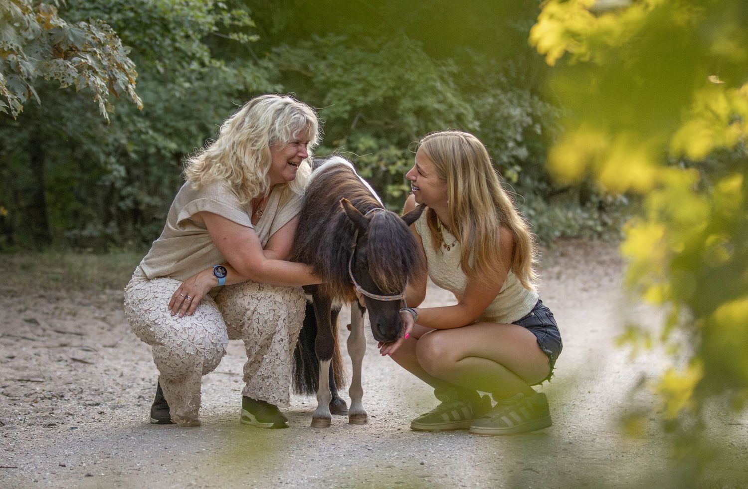 Two blonde women crouching down to pet a small brown and white pony on a sandy forest path.