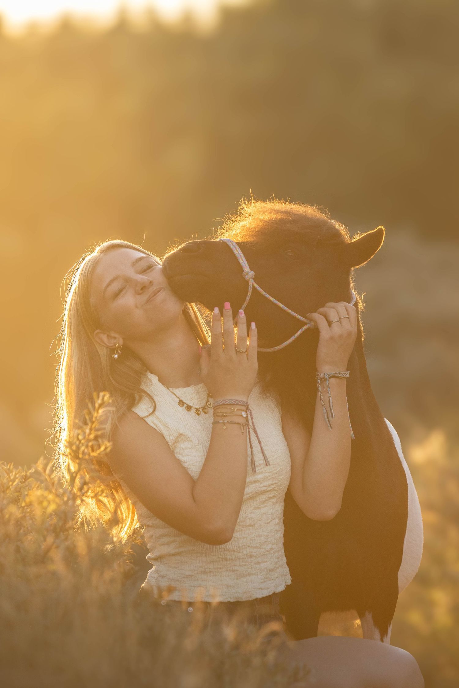 Blonde woman kissing a small dark pony bathed in warm golden sunset light in an open field.