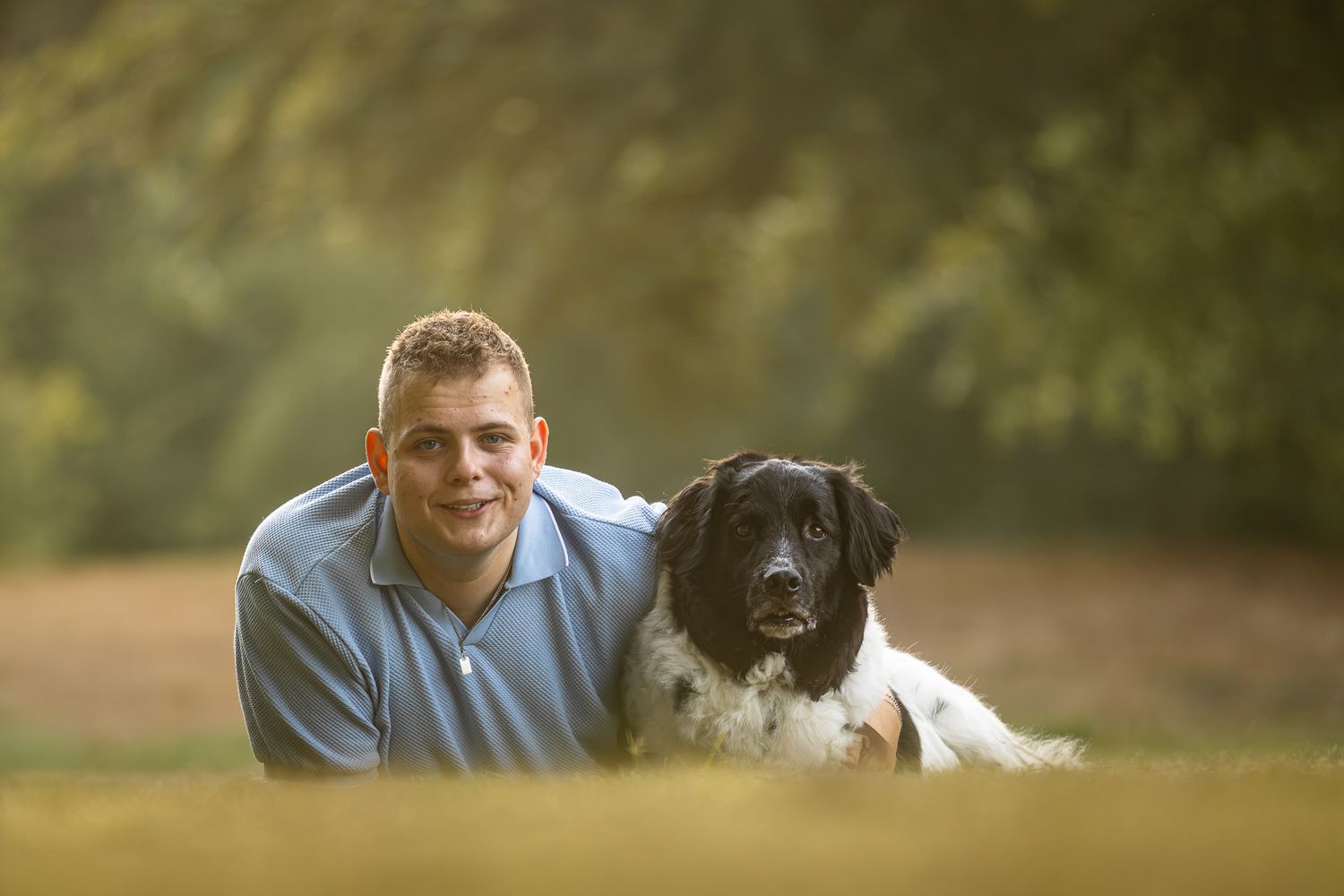 Young man posing outdoors with a black and white dog in a sunny green park setting.