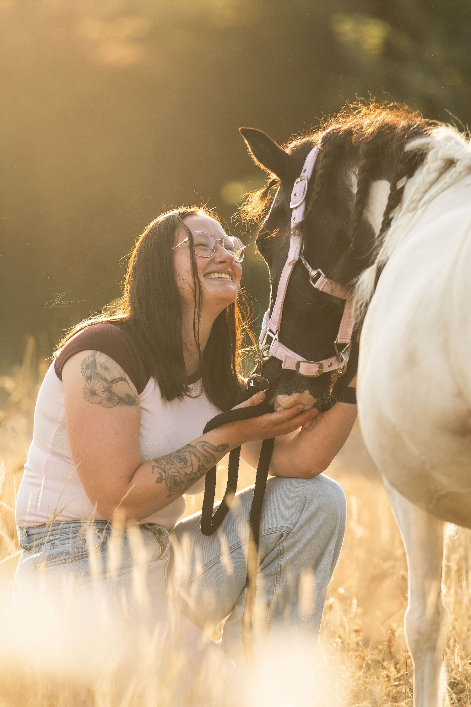 Woman with tattoos smiling joyfully while kneeling beside a horse in a sunlit golden field.