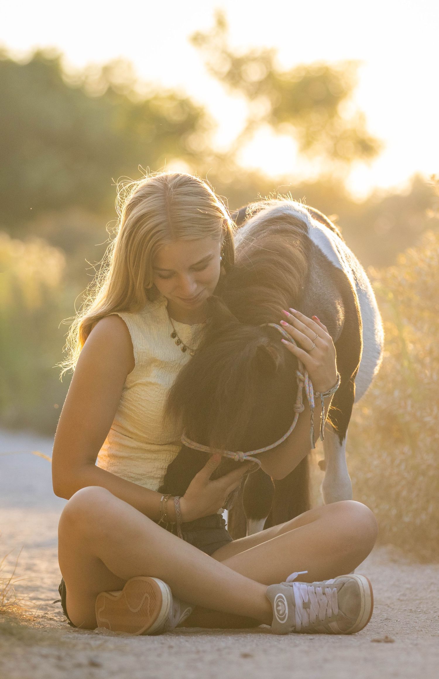 Young blonde girl sitting cross-legged on a dirt path hugging a small dark horse at golden sunset.