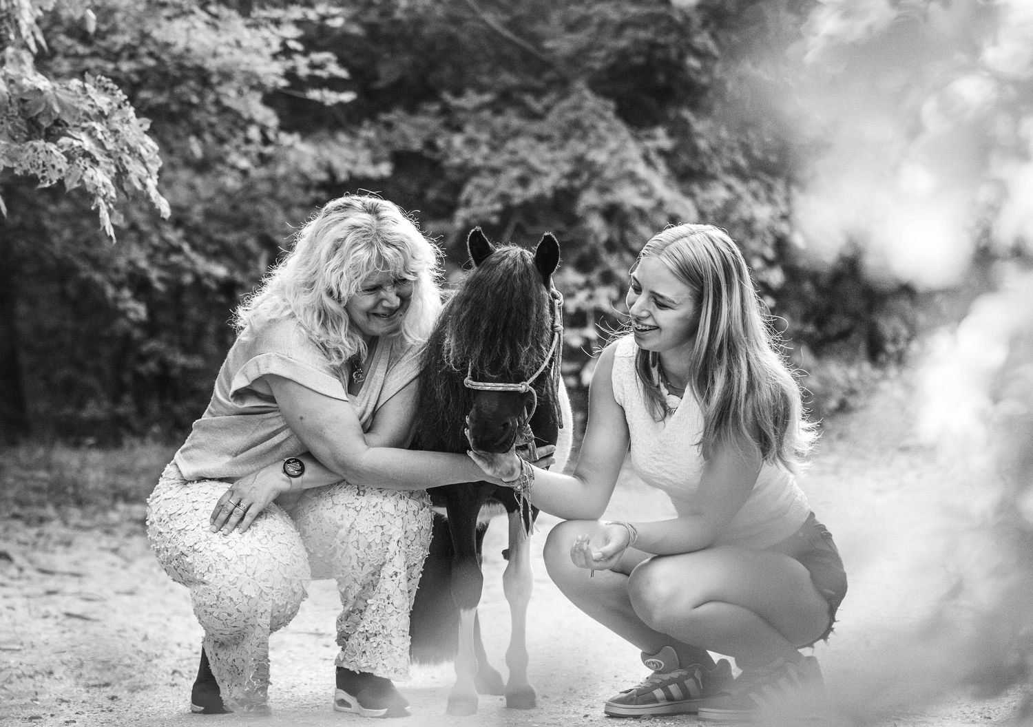 Two women crouching beside a small black pony in a forest setting, black and white photo.