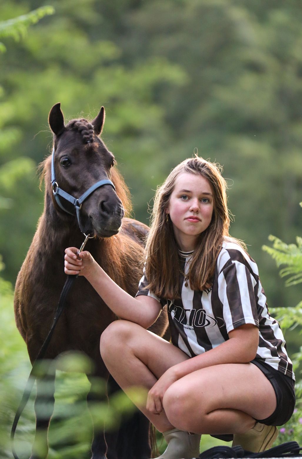 Young girl in striped shirt crouching next to a dark brown pony with a blue halter in a forest setting.