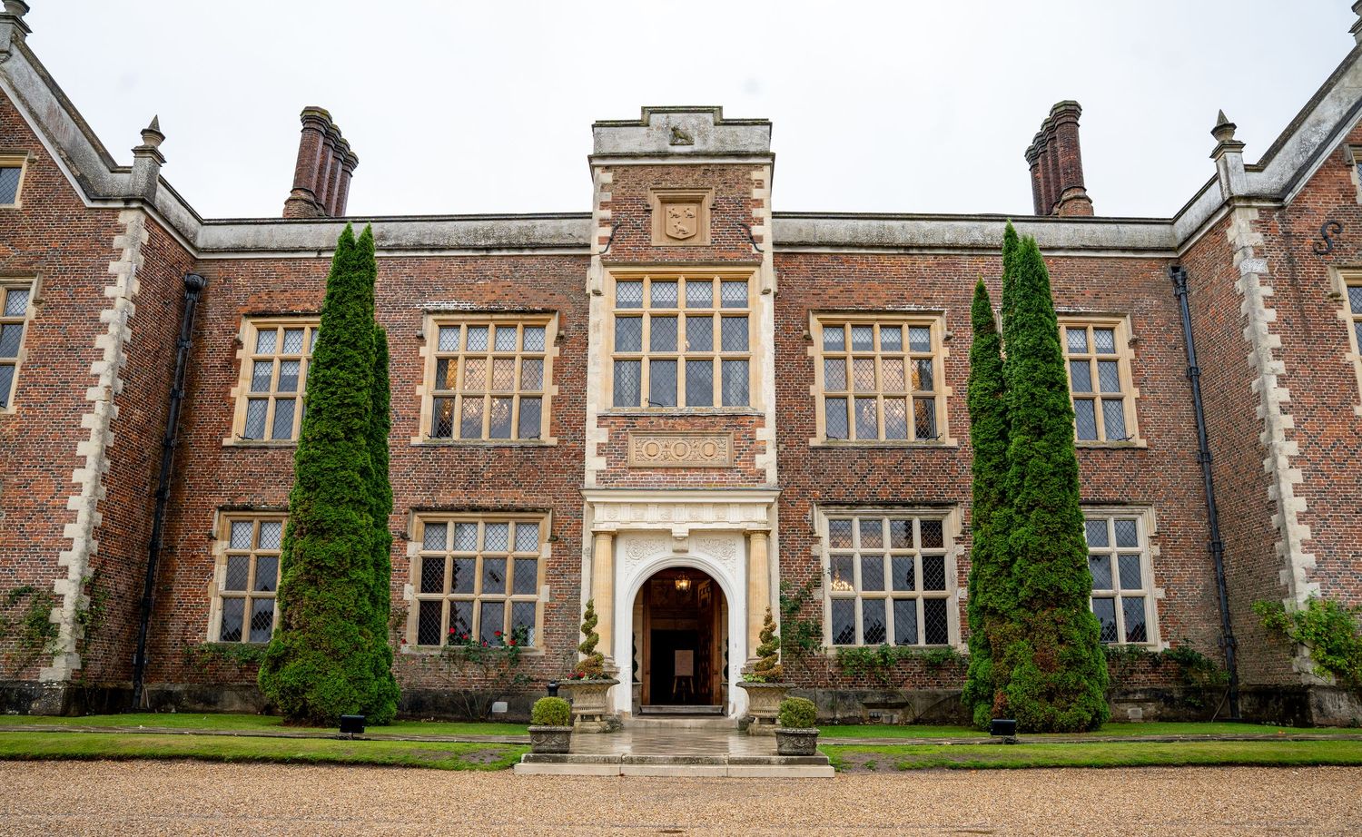Grand historic brick manor house with tall cypress trees flanking an ornate entrance doorway, set against a manicured lawn.