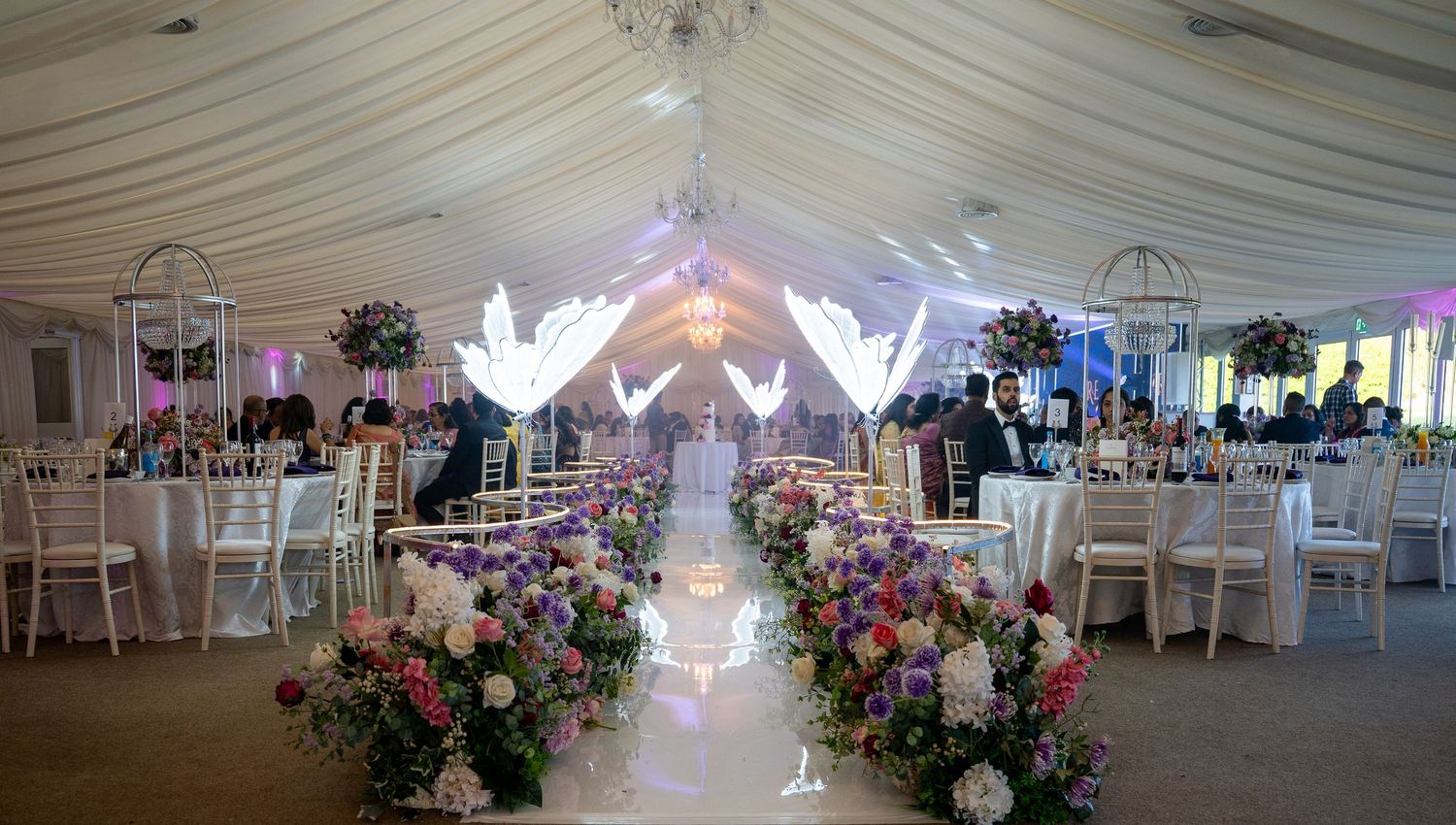 Elegant wedding reception in a white marquee tent with floral arrangements, glowing angel decor, and white chiavari chairs.