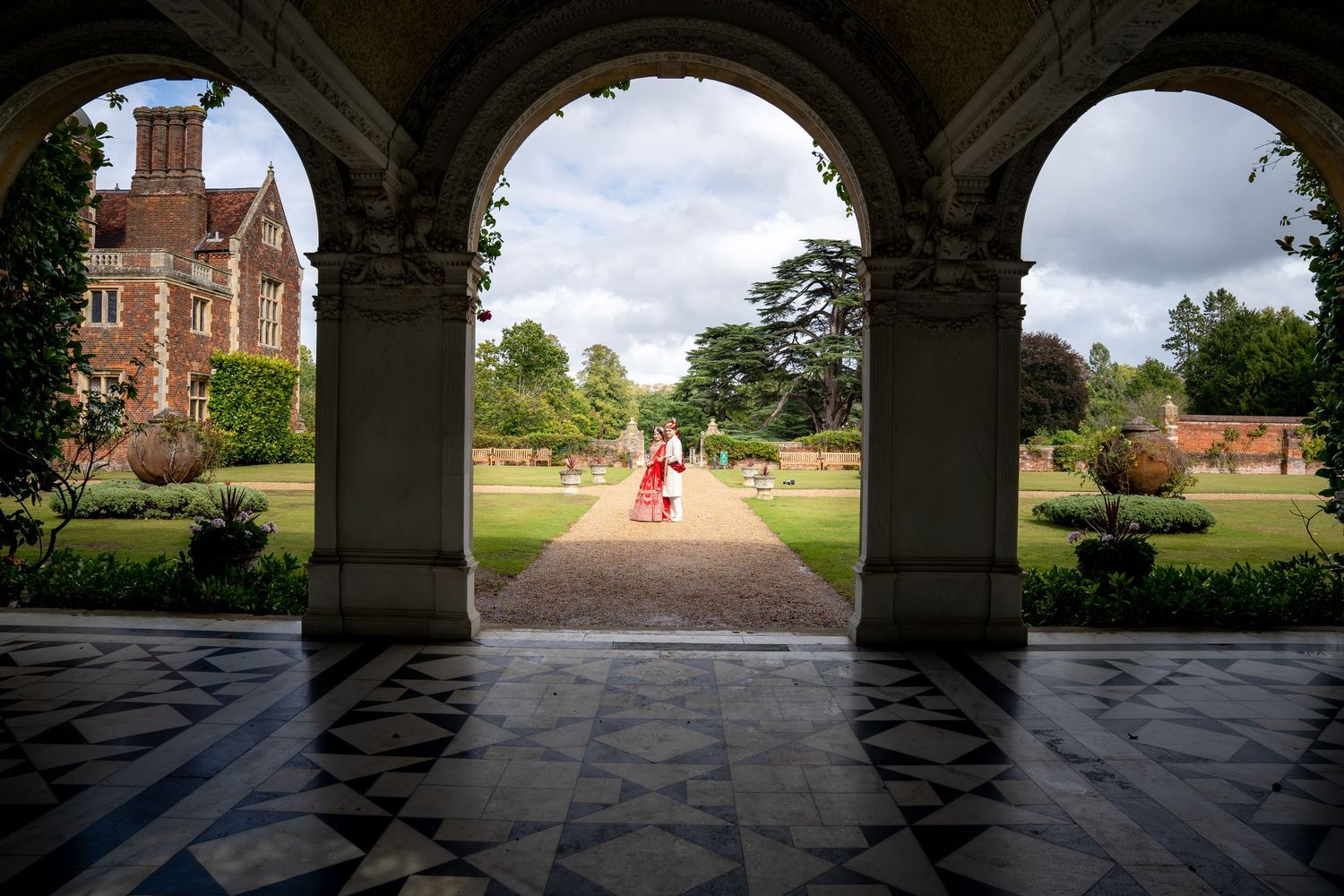 Couple walking through elegant stone archways into a formal English garden at a historic manor house.