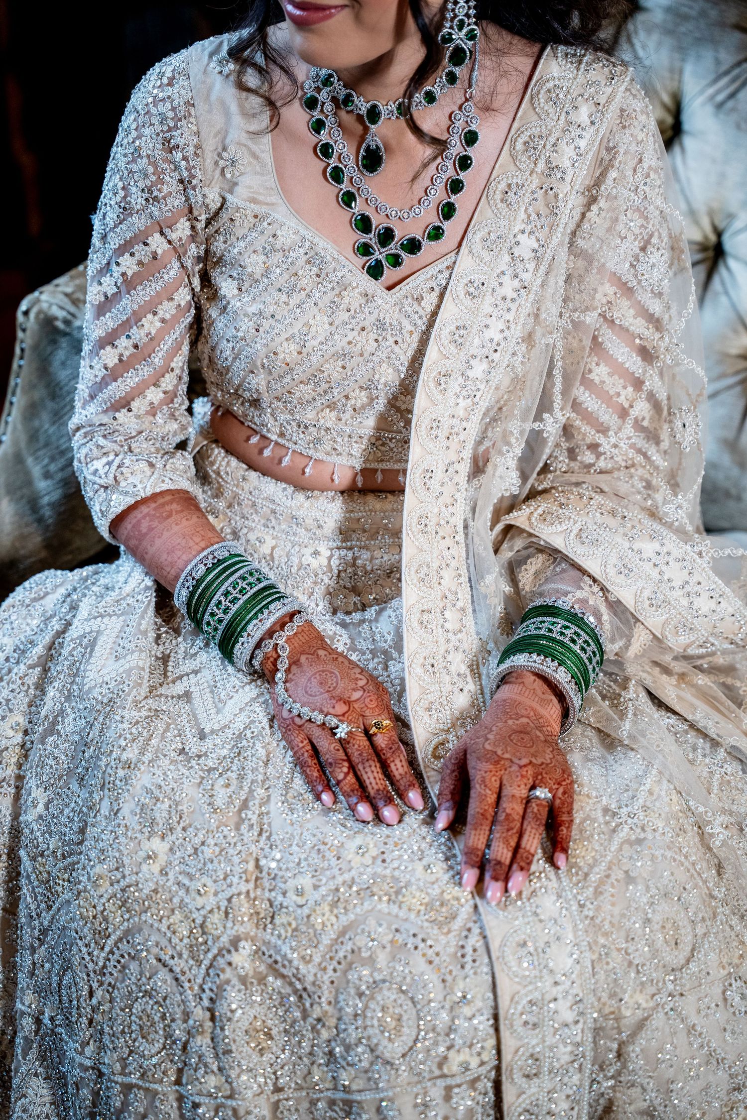 Indian bride in white lehenga displays henna hands with green bangles and emerald necklace.