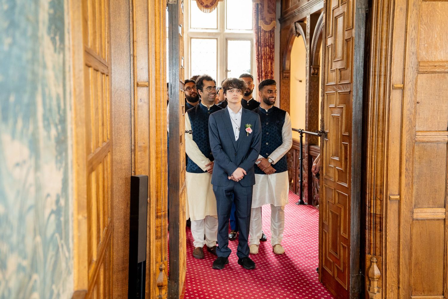 Young boy in navy suit leads group of men in traditional shalwar kameez through ornate doorway.