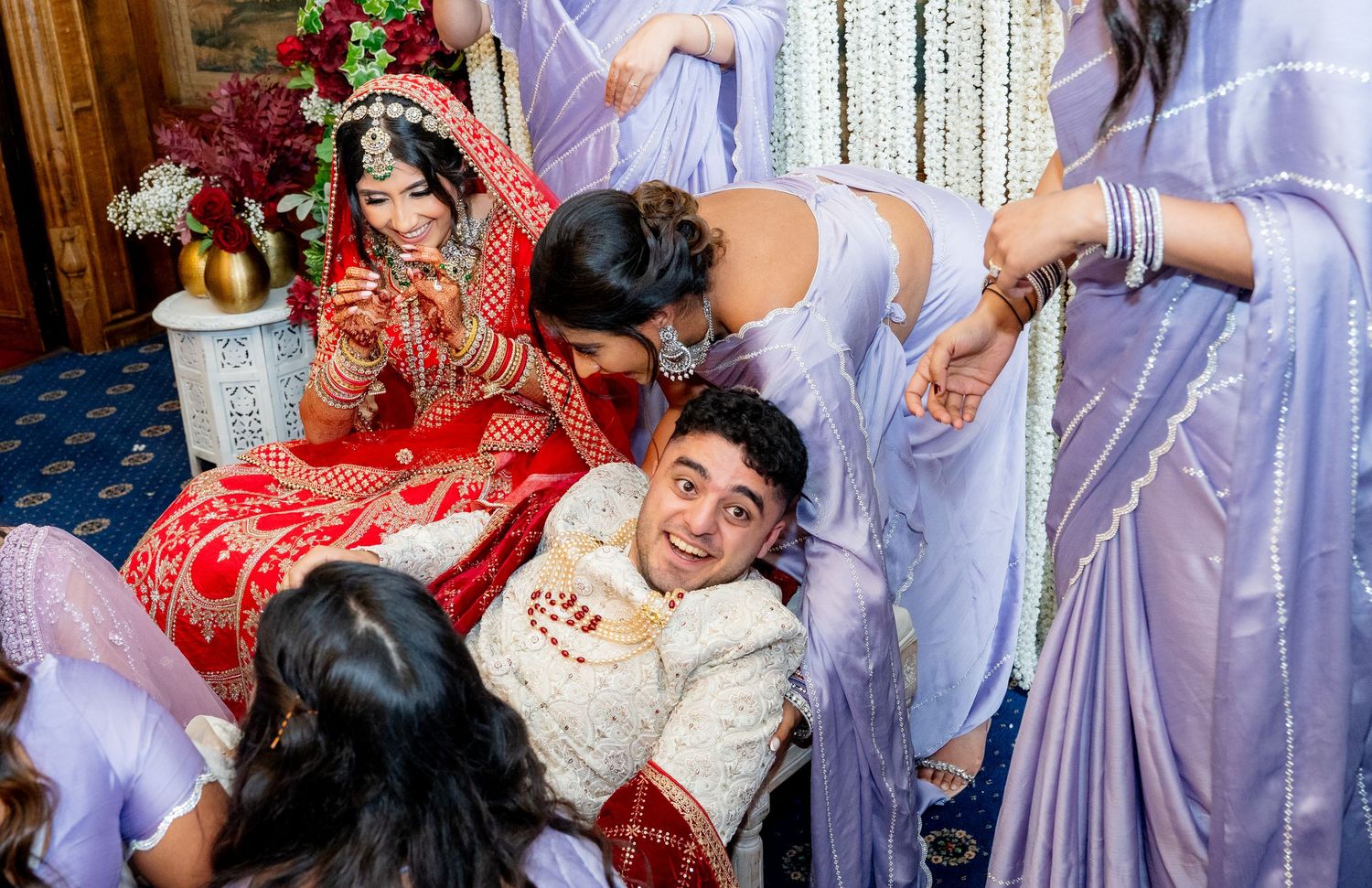 Joyful South Asian wedding moment as bridesmaids in lavender surround laughing bride and groom.