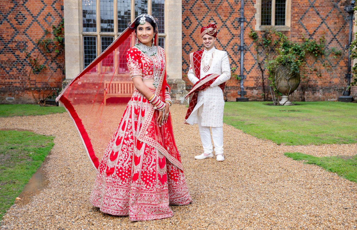 Indian bride twirling her red embroidered lehenga with groom in white sherwani outside a stately brick building.