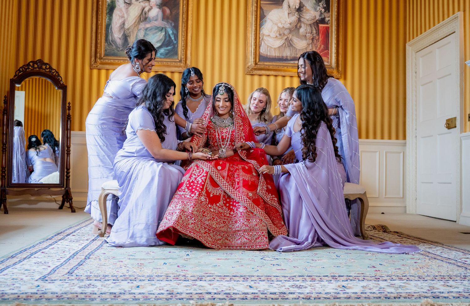 Indian bride in red lehenga surrounded by bridesmaids in lavender in an elegant gold-walled room.