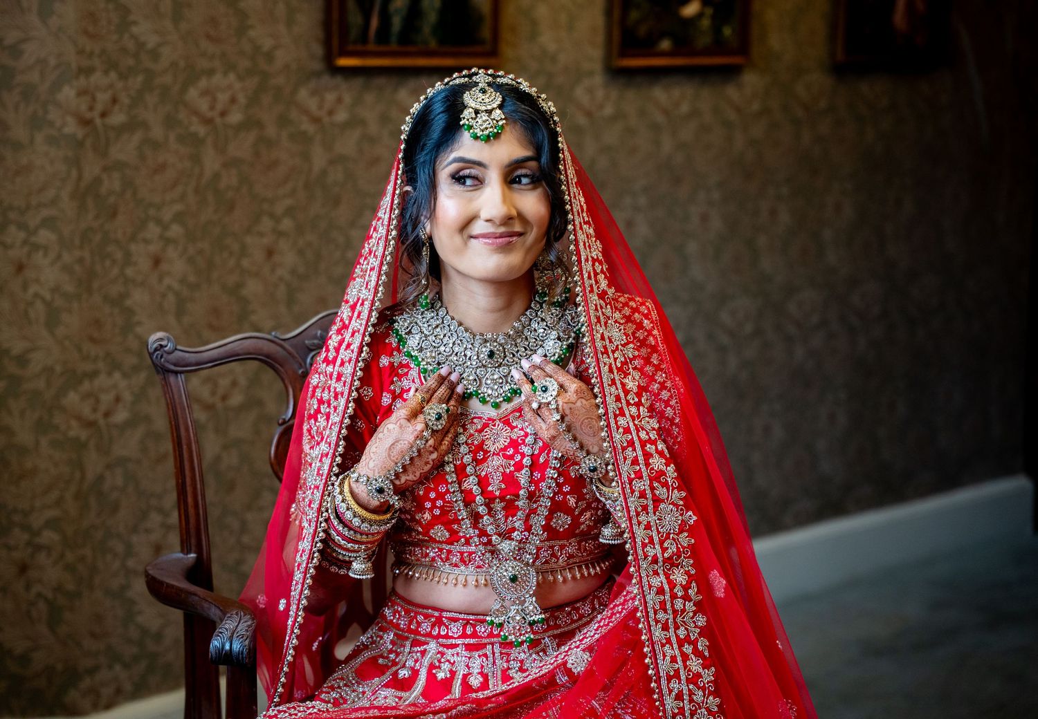 Indian bride in red bridal lehenga and gold jewelry smiling while seated in ornate chair.
