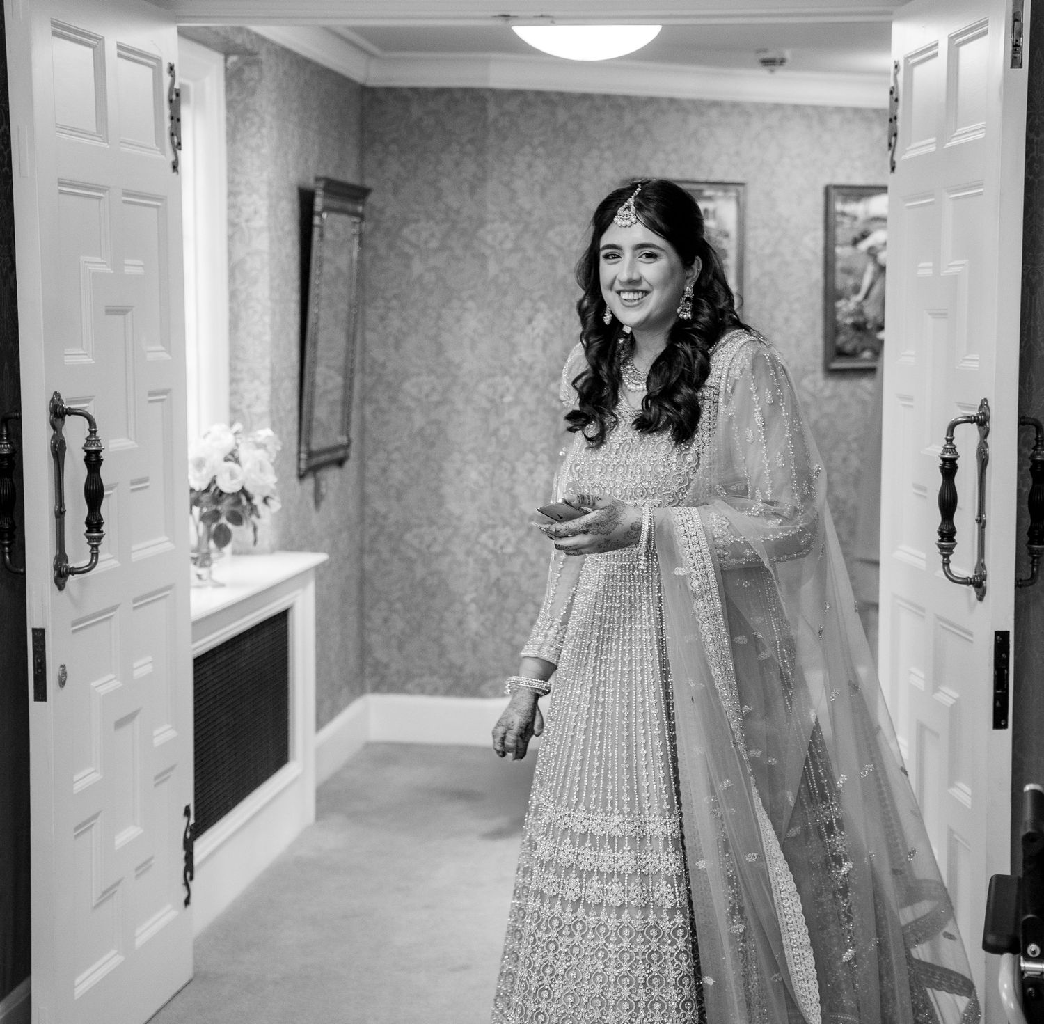Black and white photo of a smiling bride in Indian lehenga standing in a doorway.