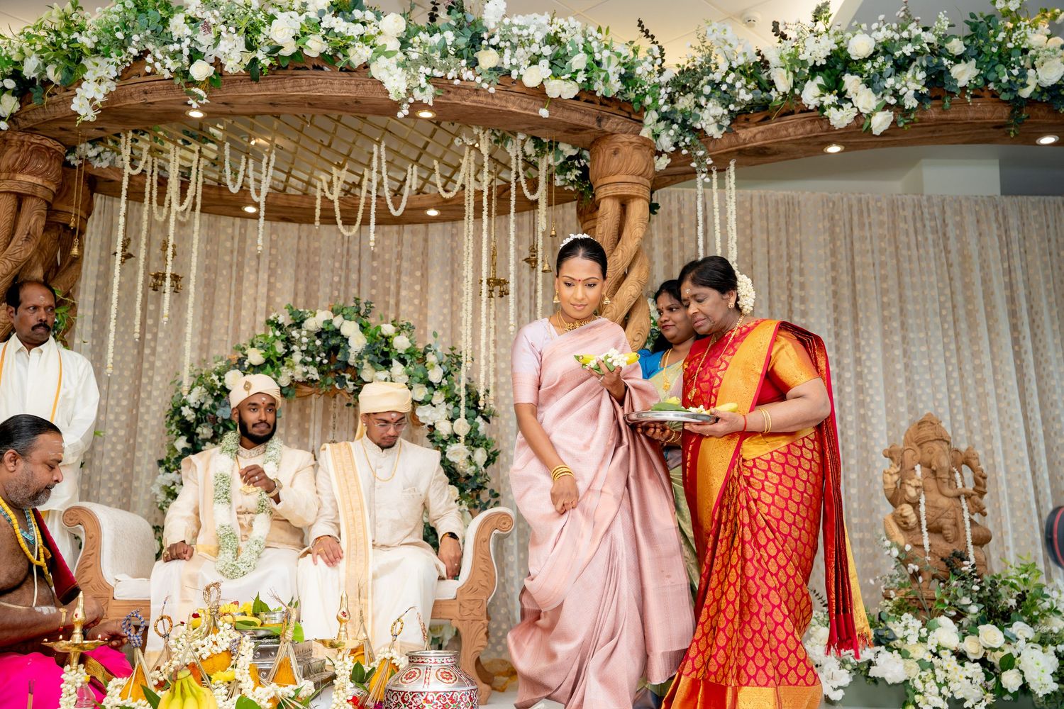Guests in traditional sarees participate in a South Indian Hindu wedding ceremony under a floral mandap.