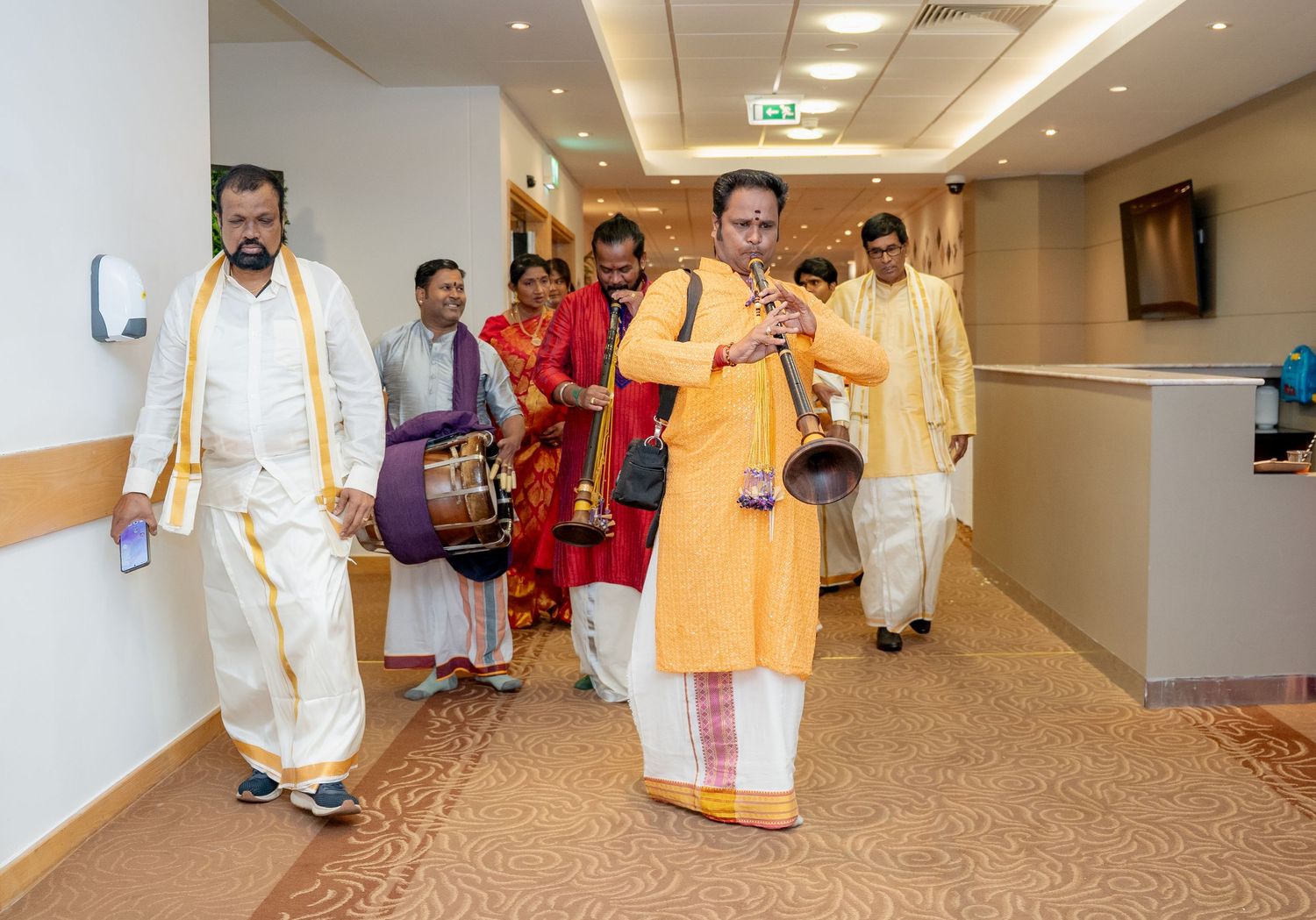 Indian wedding procession with musicians in traditional attire walking through a hotel corridor.